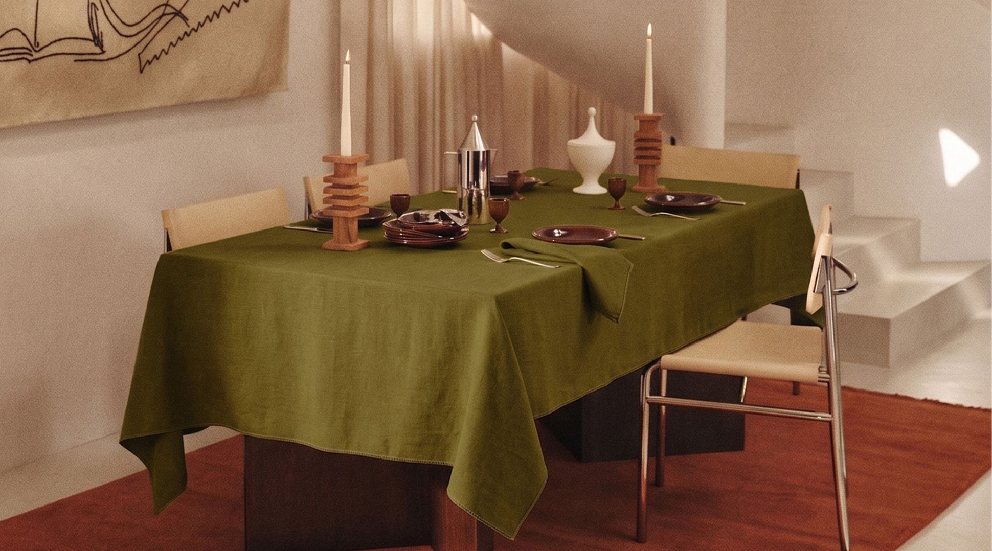 Une salle à manger moderne avec une nappe verte, décorée de bougies, de vaisselle et d'un pichet en argent. Les murs beiges sont ornés d'œuvres d'art abstrait et d'un escalier en colimaçon.