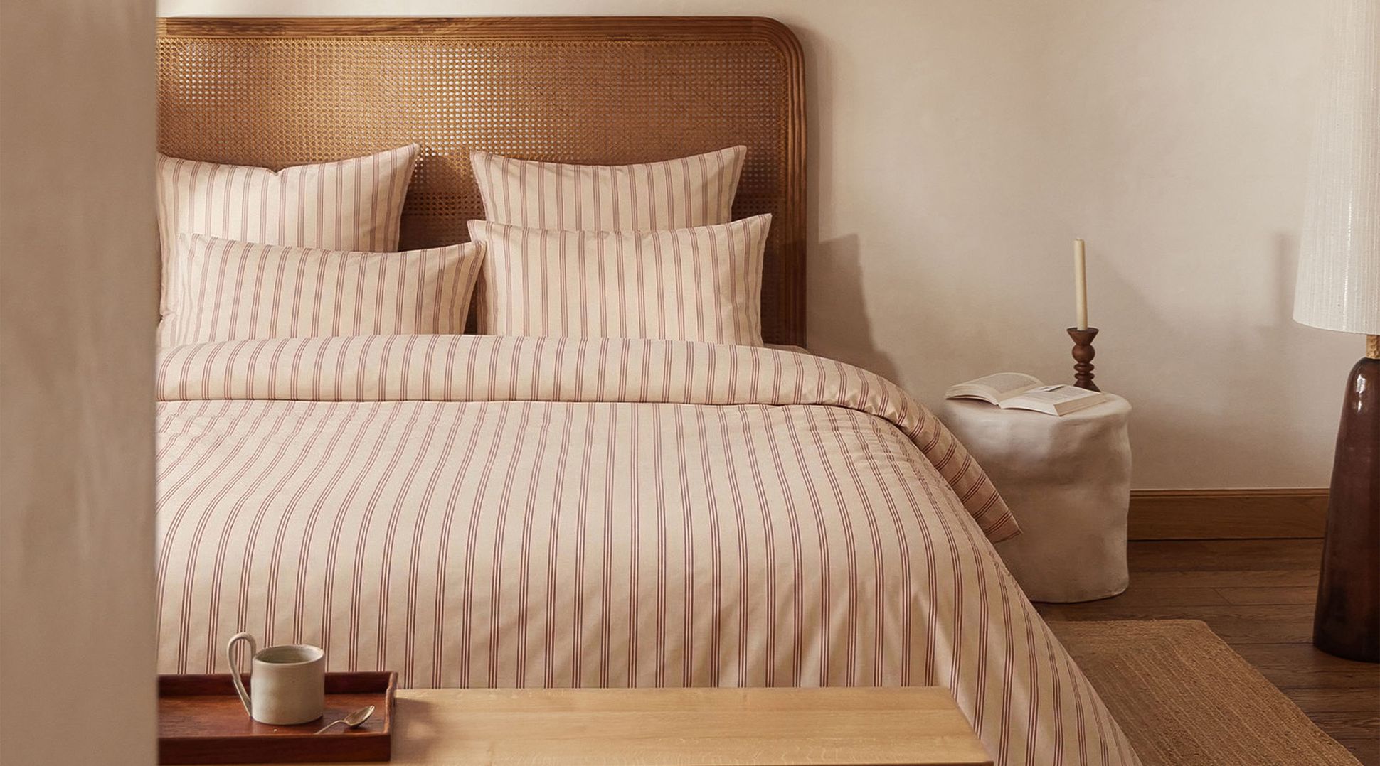 Chambre à coucher chaleureuse avec un lit orné d'une parure de lit rayée, d'une tête de lit, d'une bougie, d'un livre ouvert posé sur une petite table et d'une tasse posée sur un banc en bois.
