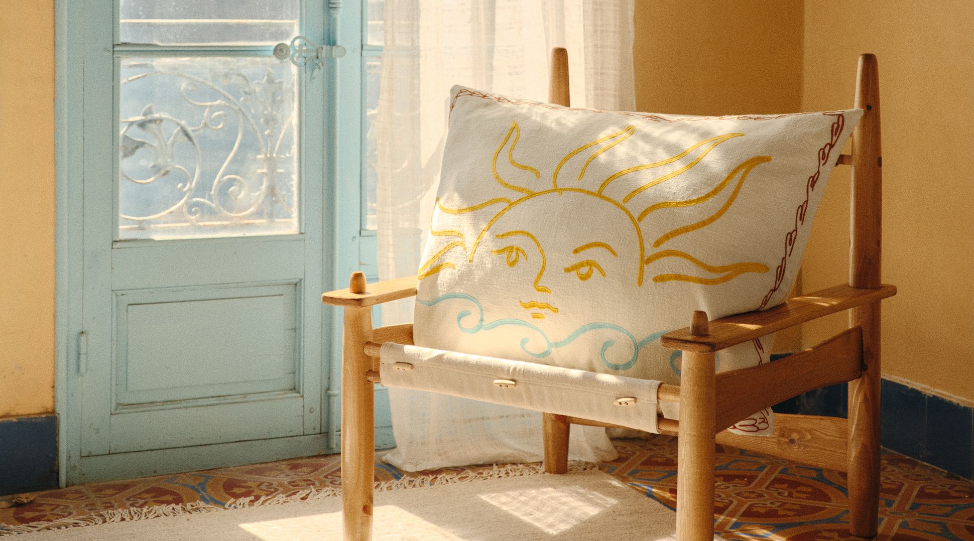 Une chaise en bois avec un coussin sur le thème du soleil, dans une pièce baignée de lumière, avec des rideaux clairs et un sol à motifs.
