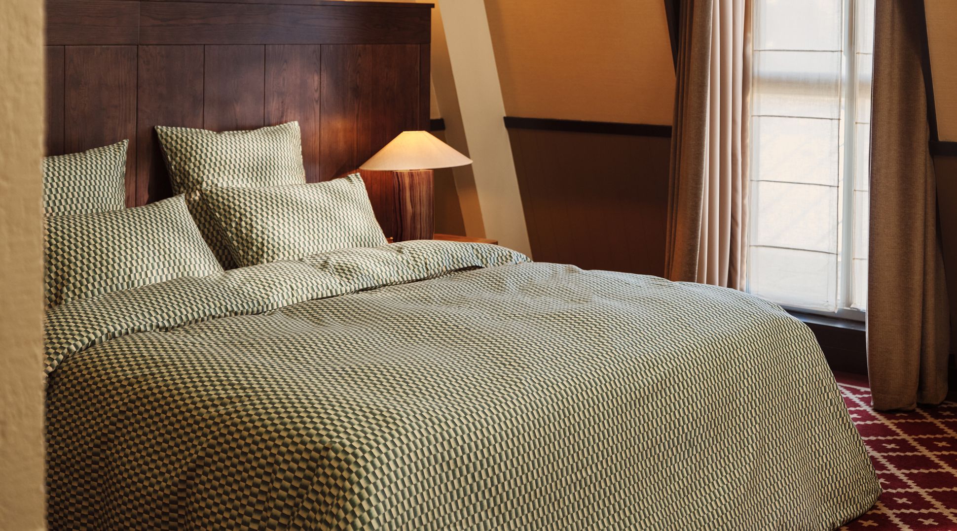 Une chambre douillette avec un lit double, une parure de lit à carreaux, une tête de lit en bois, une lampe de chevet et une fenêtre aux rideaux laissant passer une douce lumière.
