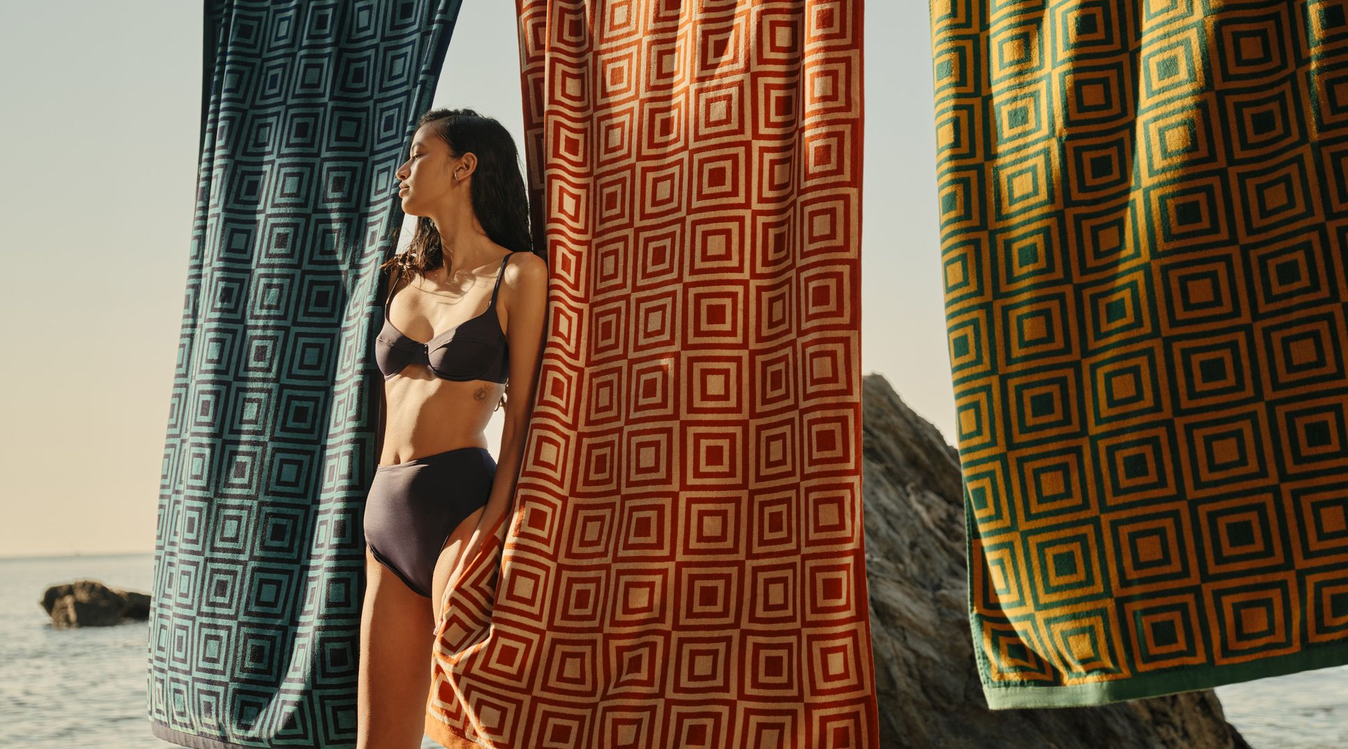 Une femme en bikini se tient au bord de la mer, encadrée par trois grandes serviettes à motifs bleues, rouges et jaunes.