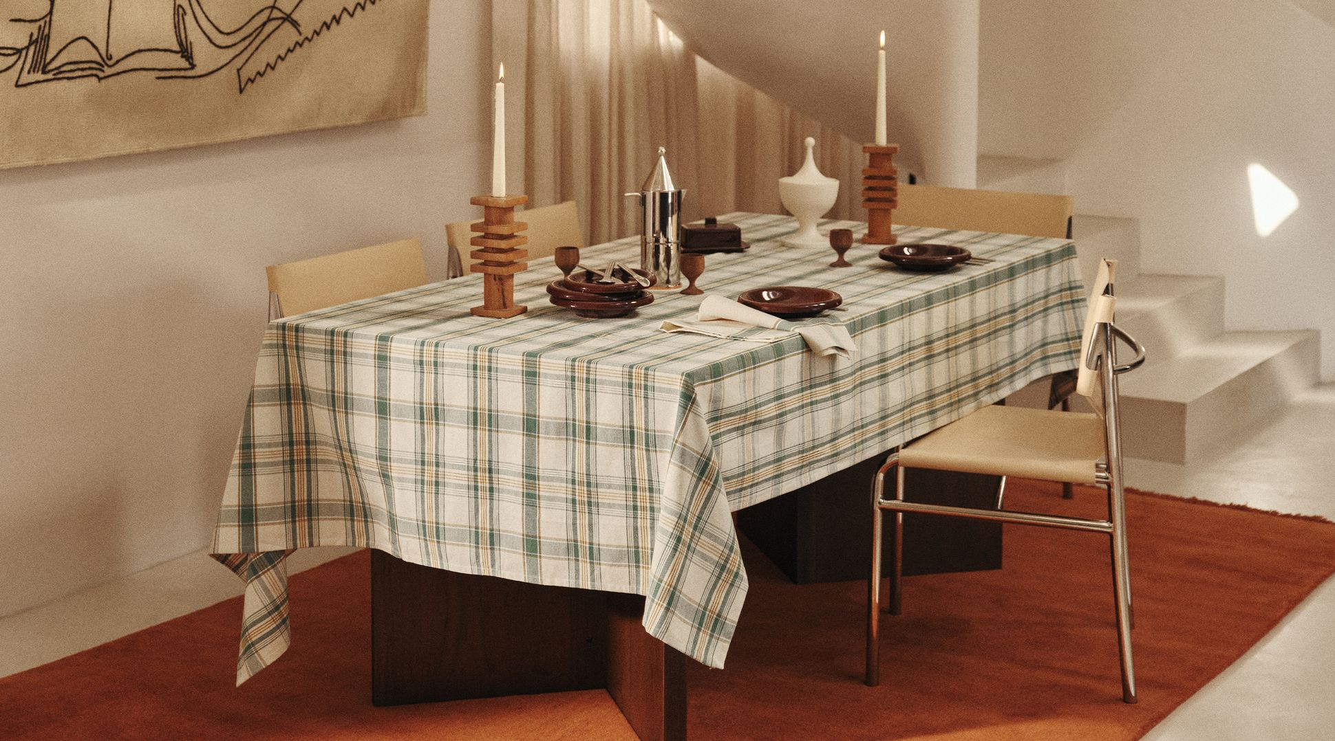 Une salle à manger avec une nappe à carreaux, dressée pour un repas avec des assiettes et des couverts, entourée de chaises, sur un tapis orange, près d'un escalier en colim