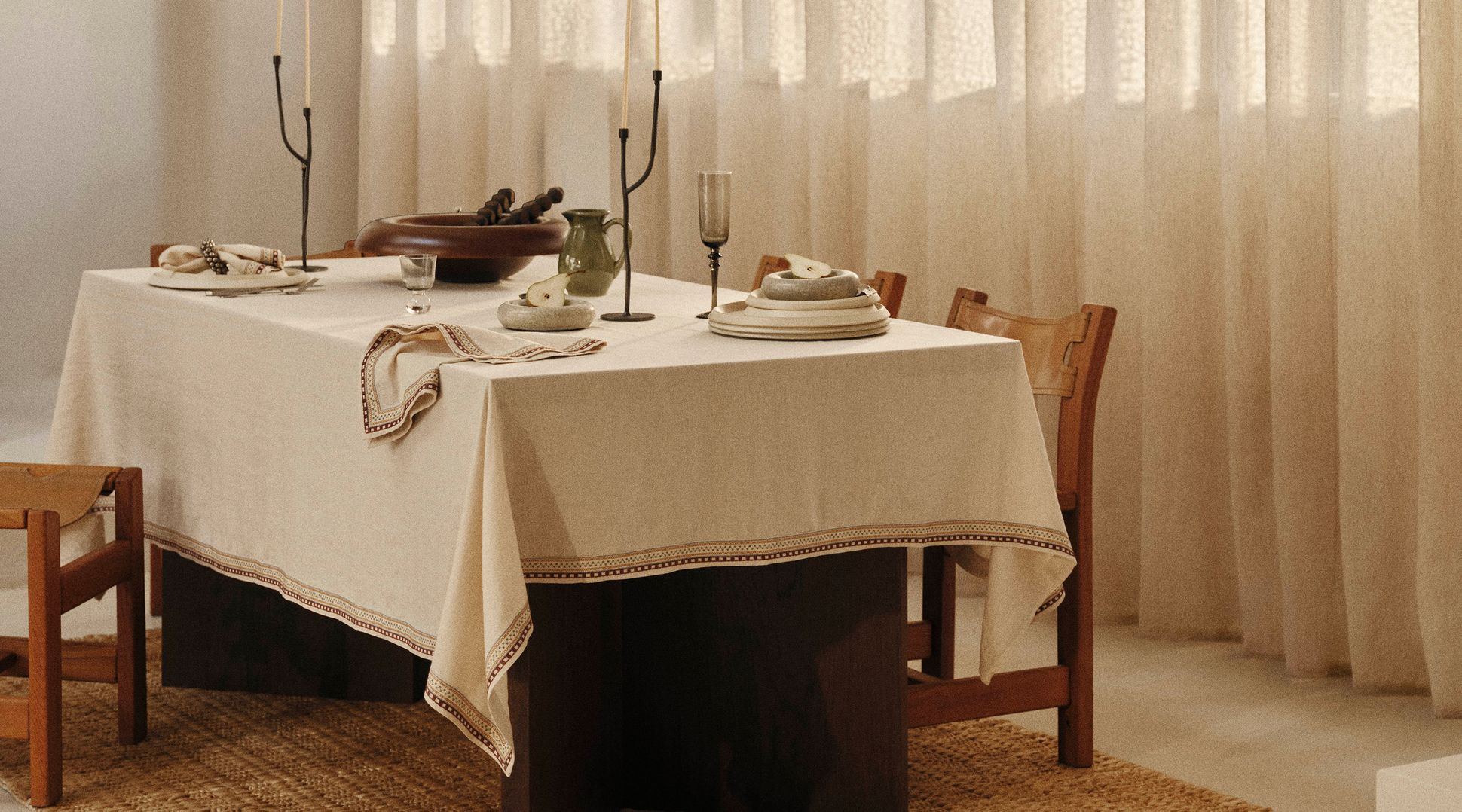Une table à manger dressée avec une nappe crème, des assiettes, des bougies, des verres et des bols en bois, entourée de chaises en bois, dans une pièce baignée d'une lumière tamisée.