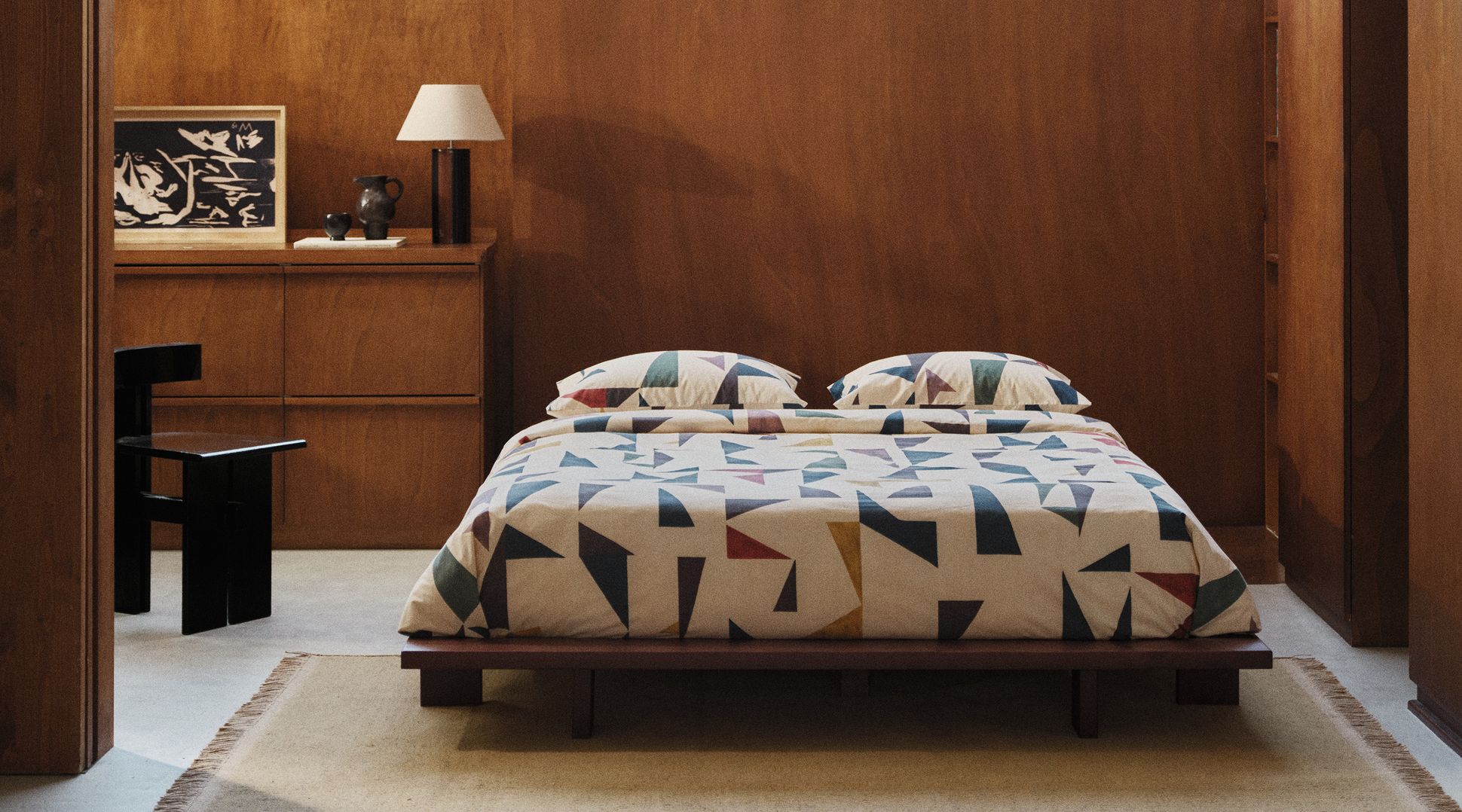 Une chambre douillette avec un lit bas recouvert d'une parure aux motifs géométriques colorés, des murs en bois et de grandes fenêtres donnant sur les arbres à l'extérieur.