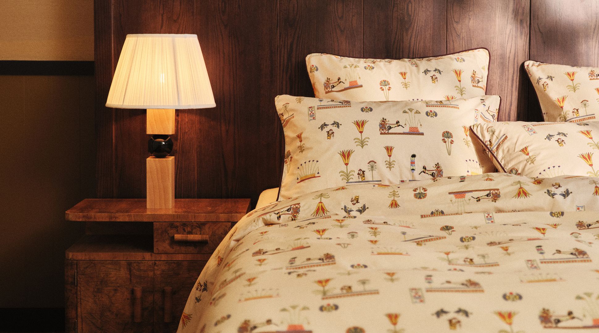 Une chambre douillette équipée d'une table de chevet en bois avec une lampe et d'un lit orné d'une parure de lit beige et d'oreillers aux motifs colorés et raffinés.