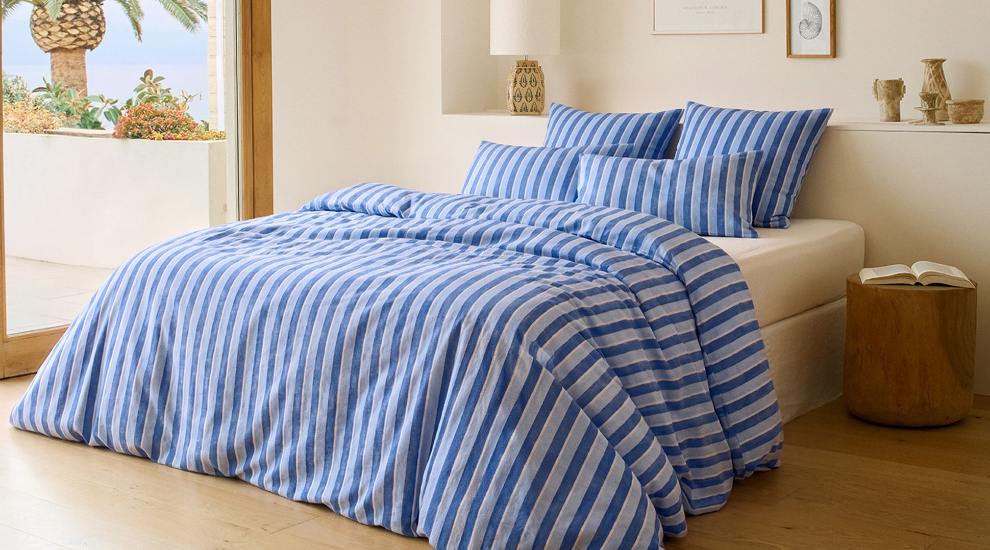 Un lit avec une parure de lit à rayures bleues et blanches, entouré d'un parquet, d'une petite table sur laquelle repose un livre, et d'une fenêtre donnant sur des palmiers.