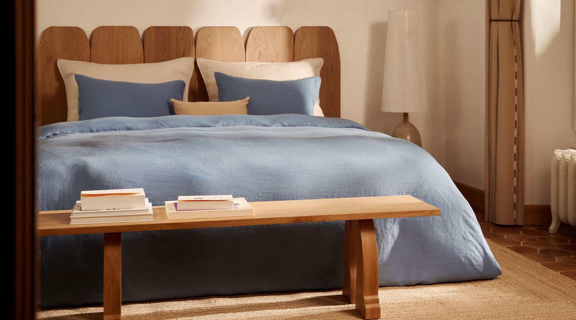 Lit douillet avec des oreillers et une couette bleu clair soigneusement disposés, une tête de lit en bois et une lampe beige dans une pièce faiblement éclairée.