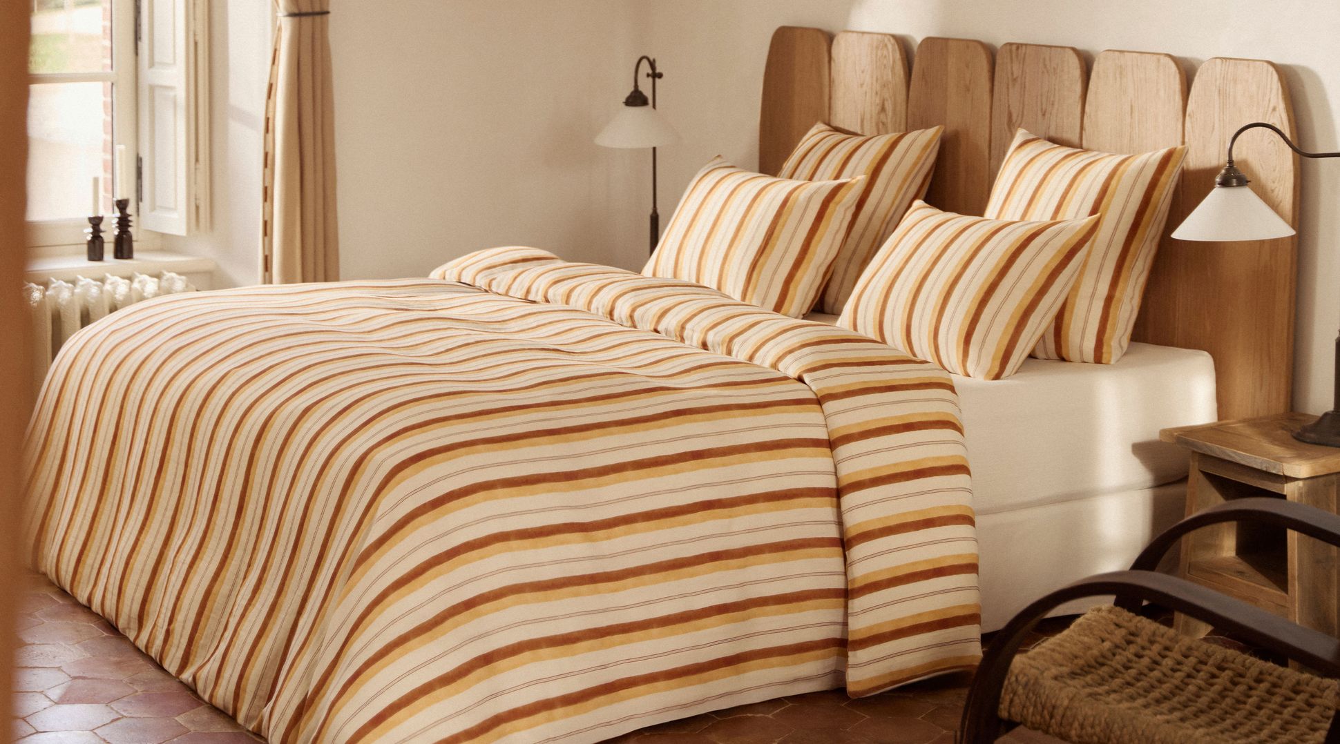 Chambre confortable avec une tête de lit en bois, une parure de lit rayée orange et beige et une décoration aux tons chauds près d'une grande fenêtre à l'éclairage doux.