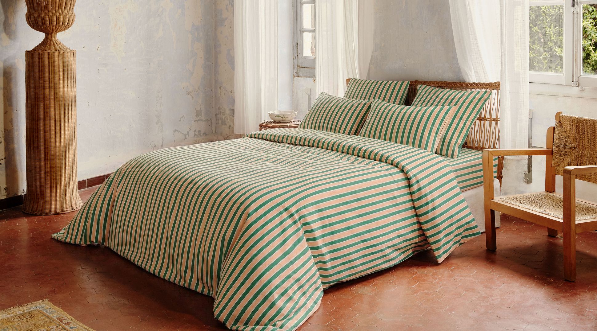 Un lit de repos en rotin avec des coussins rayés verts et beiges, à côté d'un fauteuil en bois et de rideaux blancs, dans un cadre chaleureux.