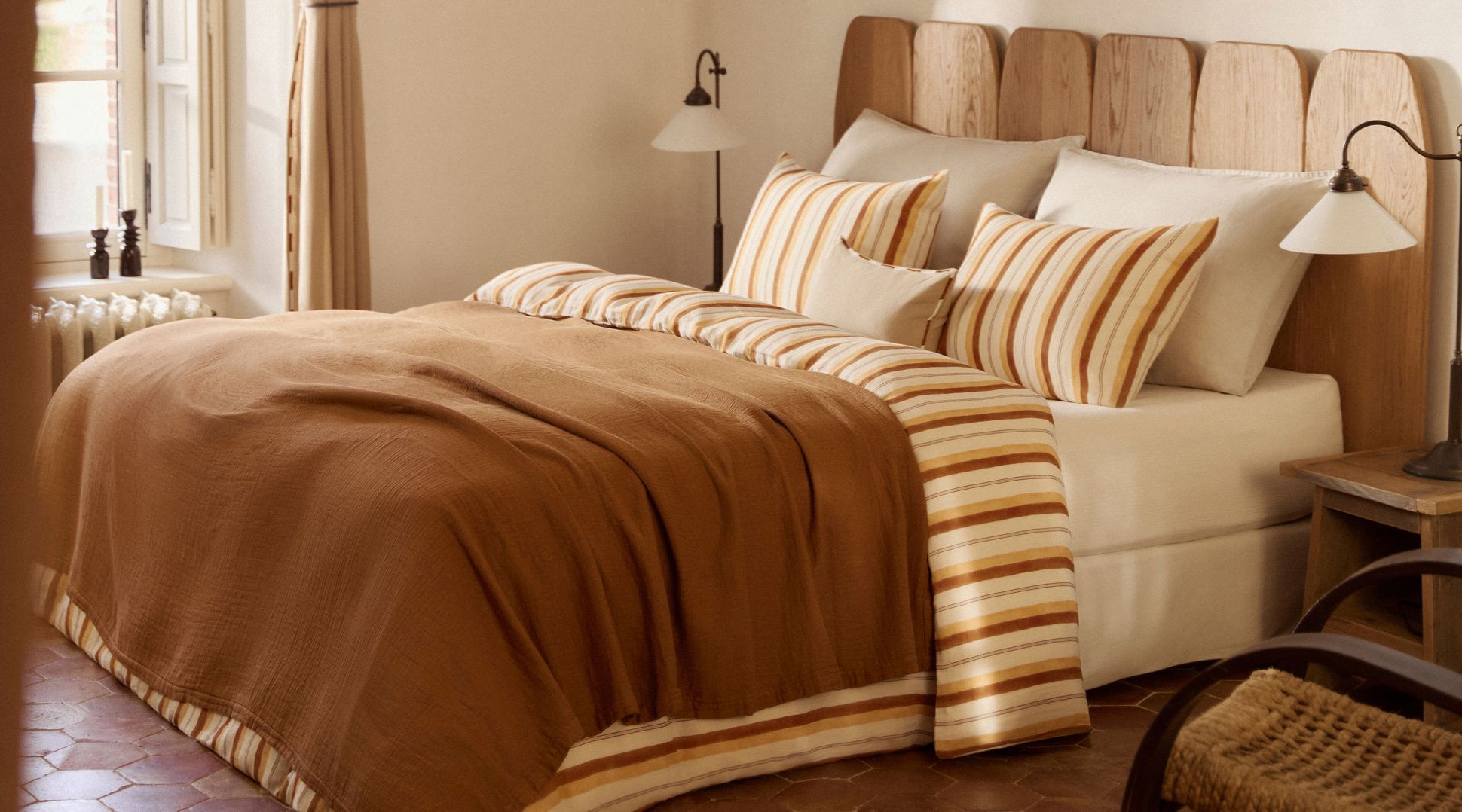 Chambre confortable avec une tête de lit en bois, des oreillers rayés et un couvre-lit marron. L'éclairage chaleureux et la lampe de chevet créent une ambiance paisible.