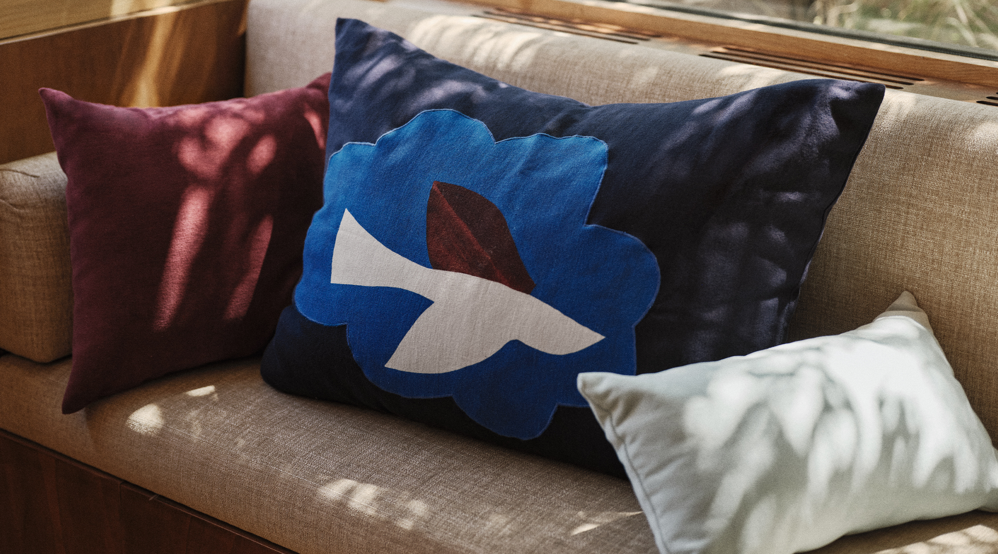 Trois coussins posés sur un canapé beige : l'un, orné d'un motif floral bleu et d'une colombe blanche, est encadré par des coussins bordeaux et vert clair, le tout baigné d'une douce lumière naturelle.