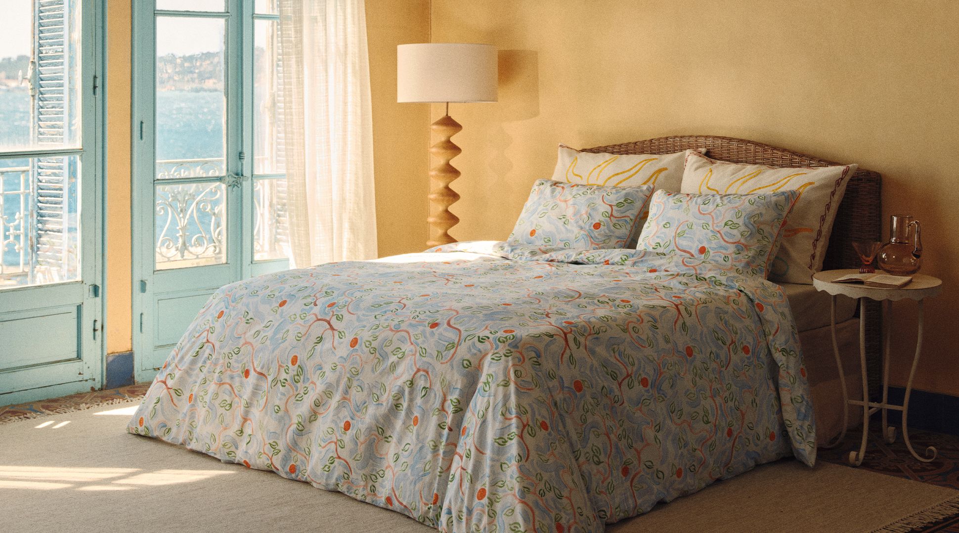Une chambre douillette avec une tête de lit en rotin, une parure de lit aux motifs floraux colorés et une lampe élégante posée contre un mur aux tons chauds.
