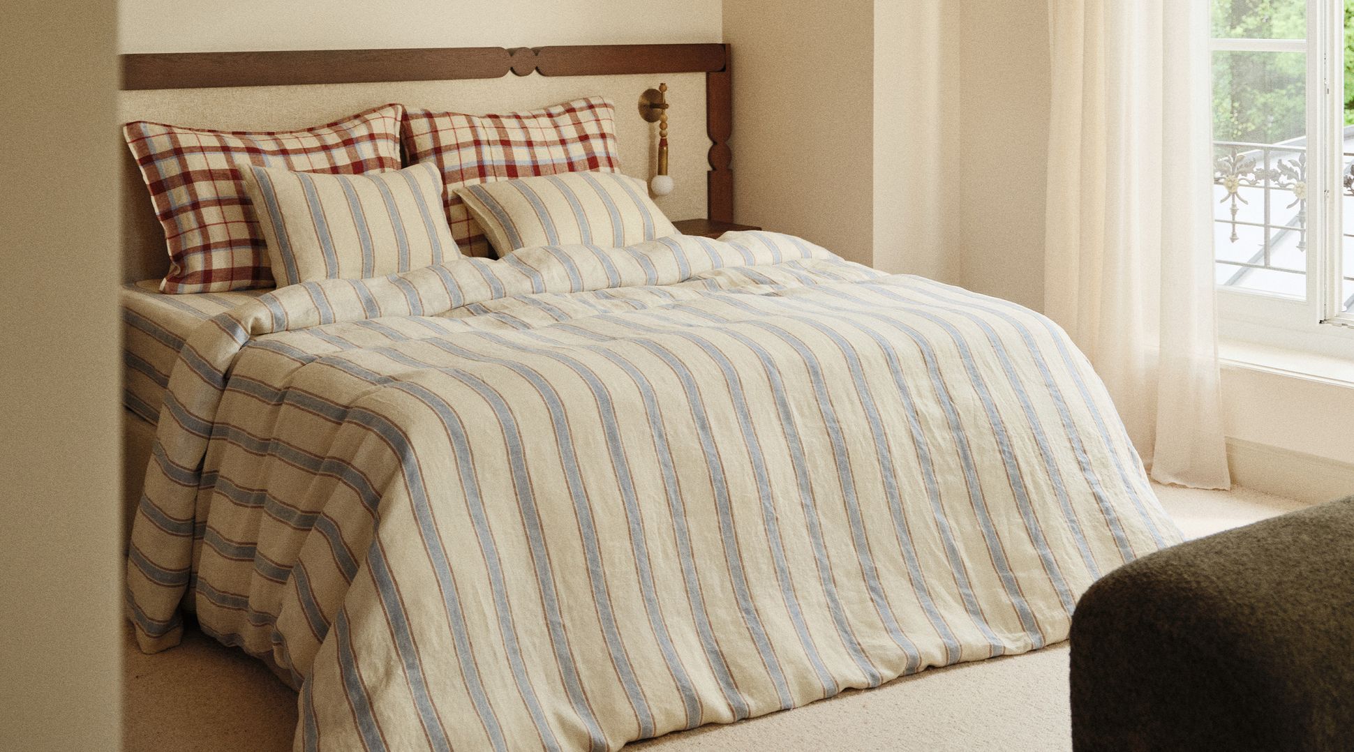 Chambre à coucher chaleureuse, avec un lit garni de draps rayés et d'oreillers à carreaux, près d'une fenêtre ornée de voilages et offrant une vue sur la verdure à l'extérieur.