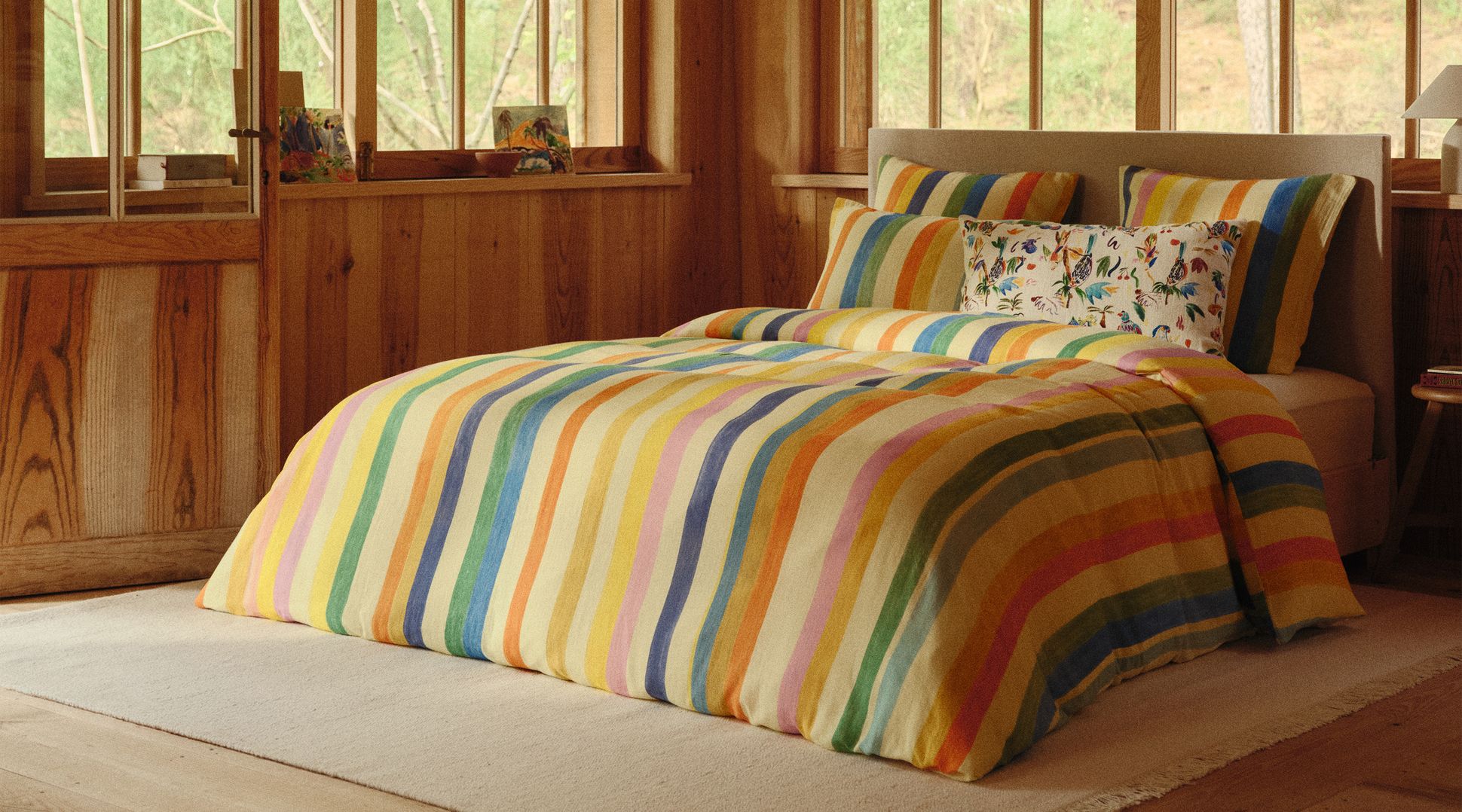 Une chambre chaleureuse en bois dotée de grandes fenêtres, avec un lit orné d'une parure de lit à rayures colorées et d'oreillers décoratifs, sous une lanterne suspendue aux couleurs vives.