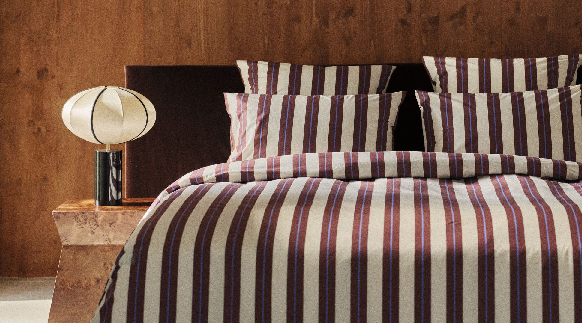 Une parure de lit rayée sur un lit, à côté d'une table de chevet en bois sur laquelle repose une lampe décorative, avec en arrière-plan un mur en bois.