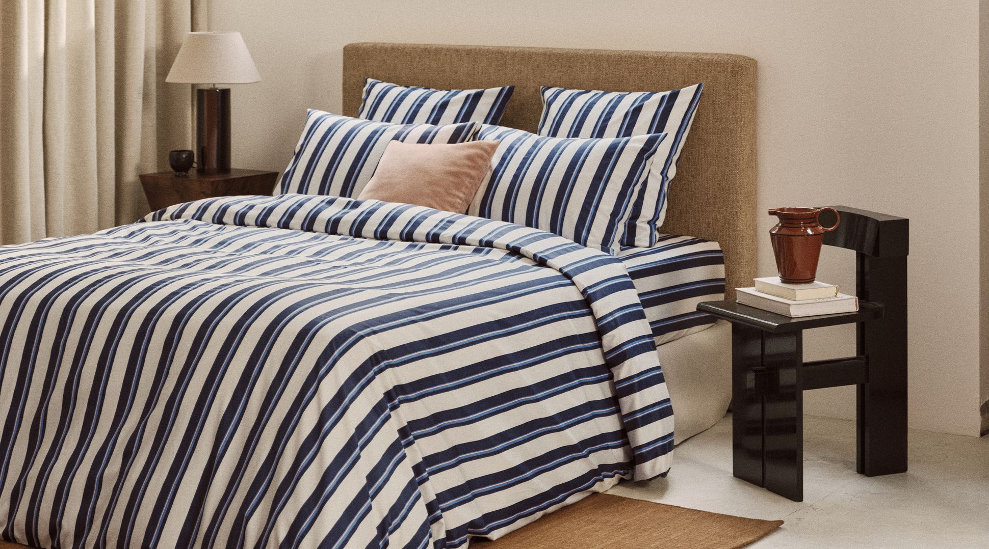 Chambre à coucher chaleureuse avec un lit habillé d'un linge de lit à rayures bleues et blanches, une tête de lit beige et une petite table de chevet sur laquelle repose une lampe, près d'une grande fenêtre.