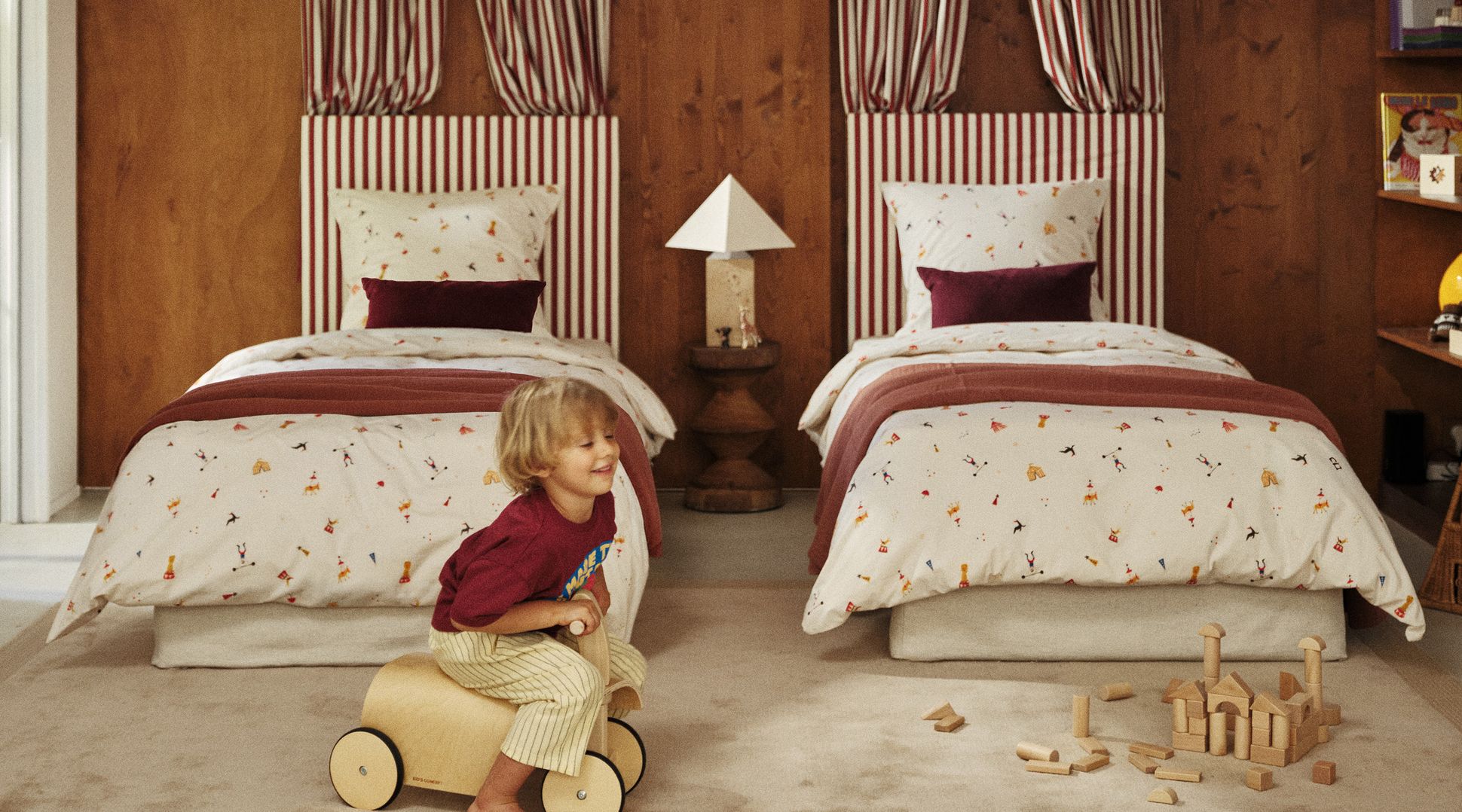 Un enfant joue avec un jouet en bois dans une chambre douillette équipée de deux lits, de rideaux rayés, d'une parure de lit aux motifs ludiques, de blocs en bois et d'une lampe posée sur une table de chevet.
