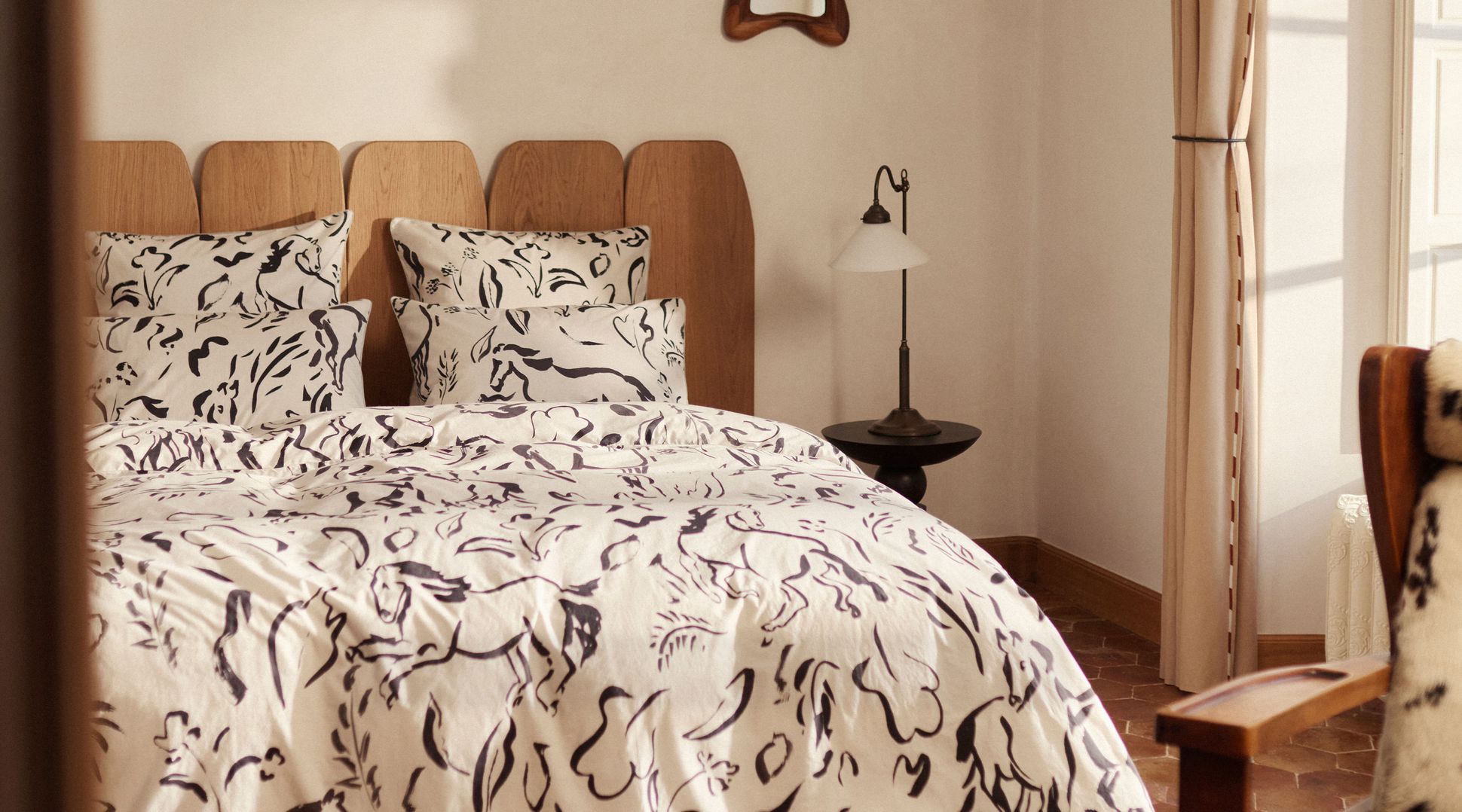 Chambre confortable avec literie blanche à motifs abstraits noirs, tête de lit en bois, petite table d'appoint avec lampe et fenêtre à rideaux.