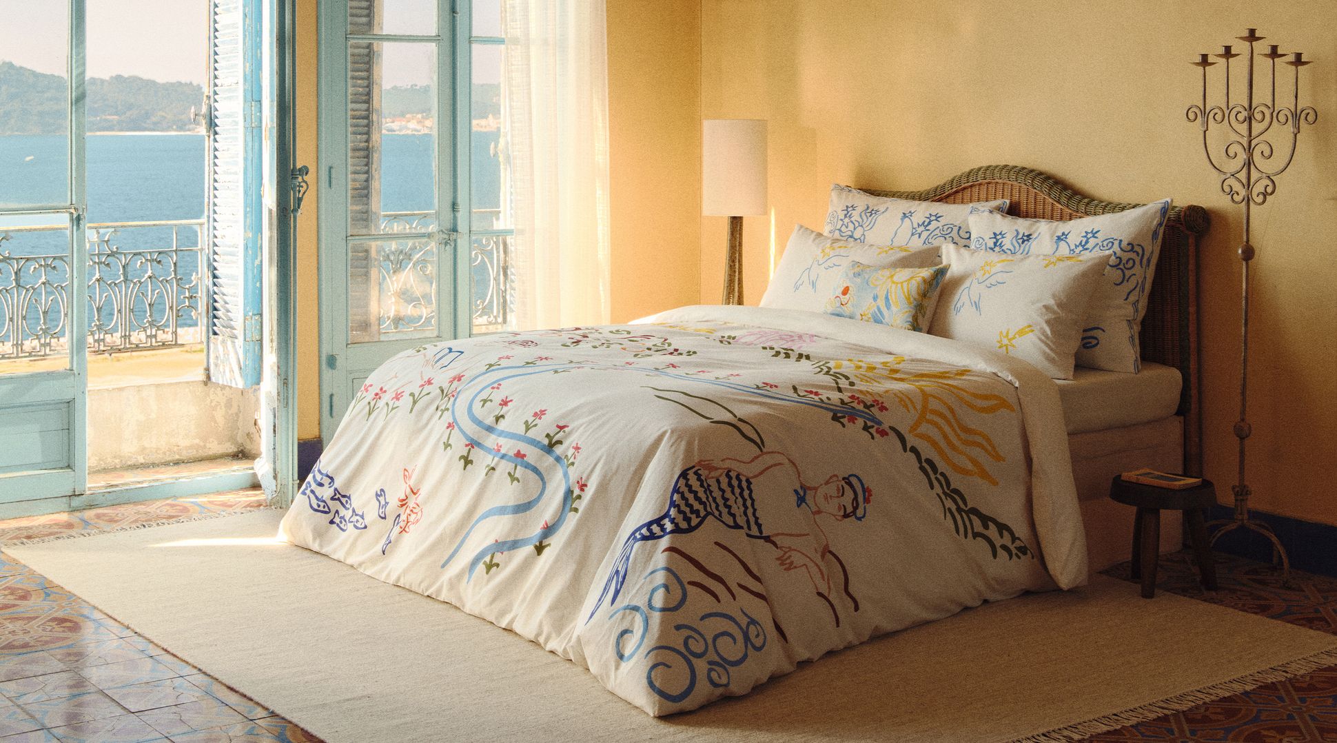 Une chambre baignée de soleil, avec une parure de lit blanche ornée de motifs abstraits colorés, une tête de lit en osier et un grand lampadaire placé près d'une fenêtre aux rideaux vaporeux.