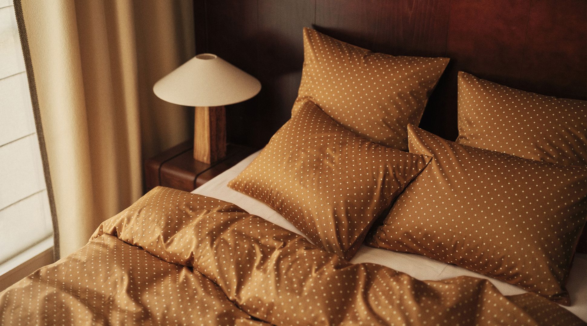 A cozy bed with brown polka dot bedding, wooden headboard, and a lamp on a bedside table, set next to beige curtains.