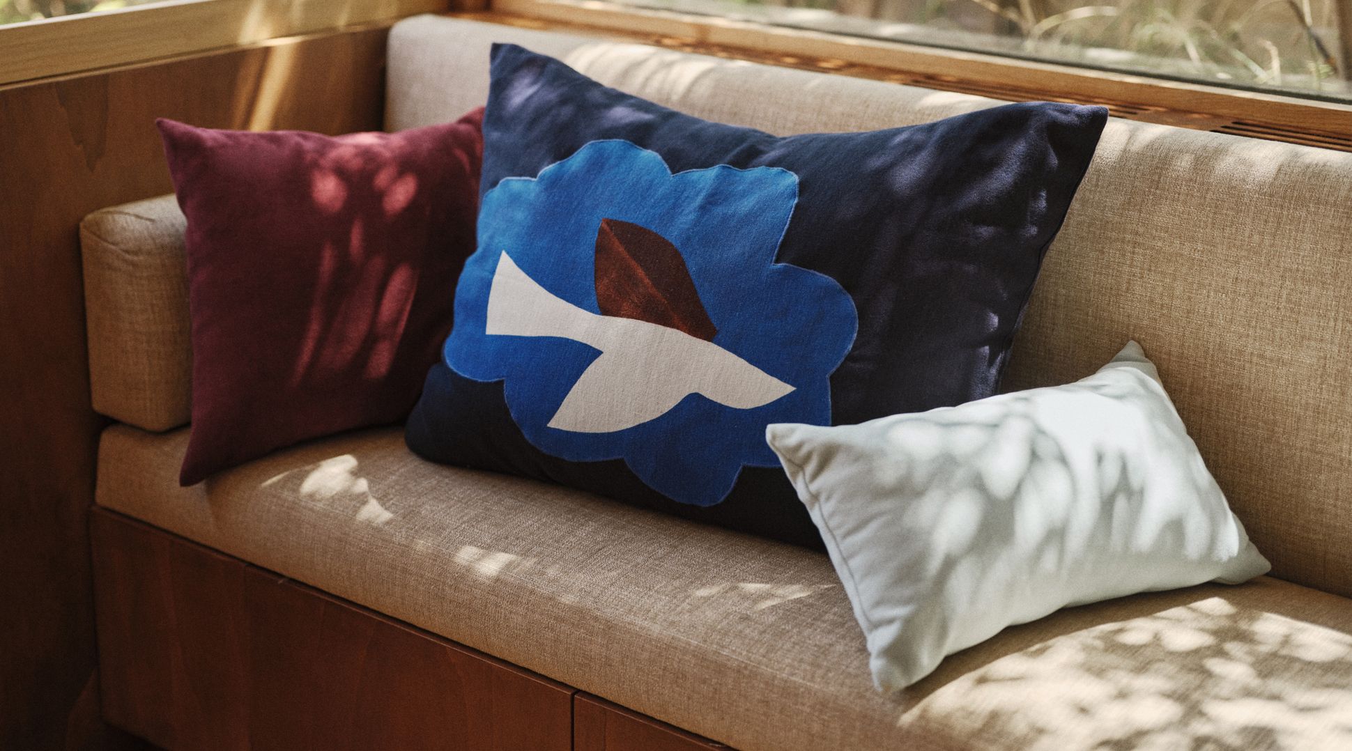 Three cushions sit on a beige sofa, one with a blue flower and bird design, and the others in burgundy and white. Sunlight filters through trees.