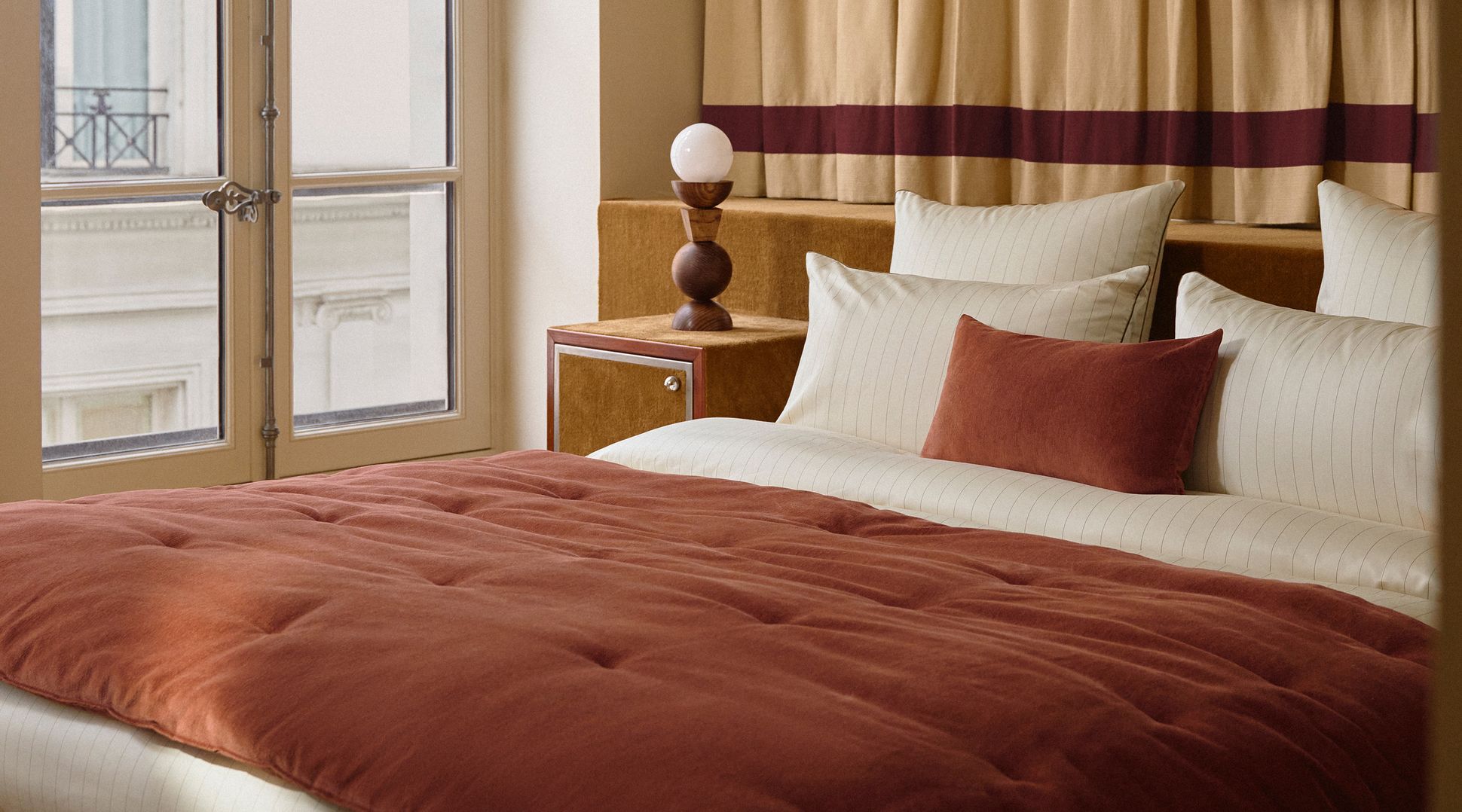 A cozy bed with rust-colored bedding and pillows next to a window. A small wooden nightstand holds a modern lamp, surrounded by beige curtains.