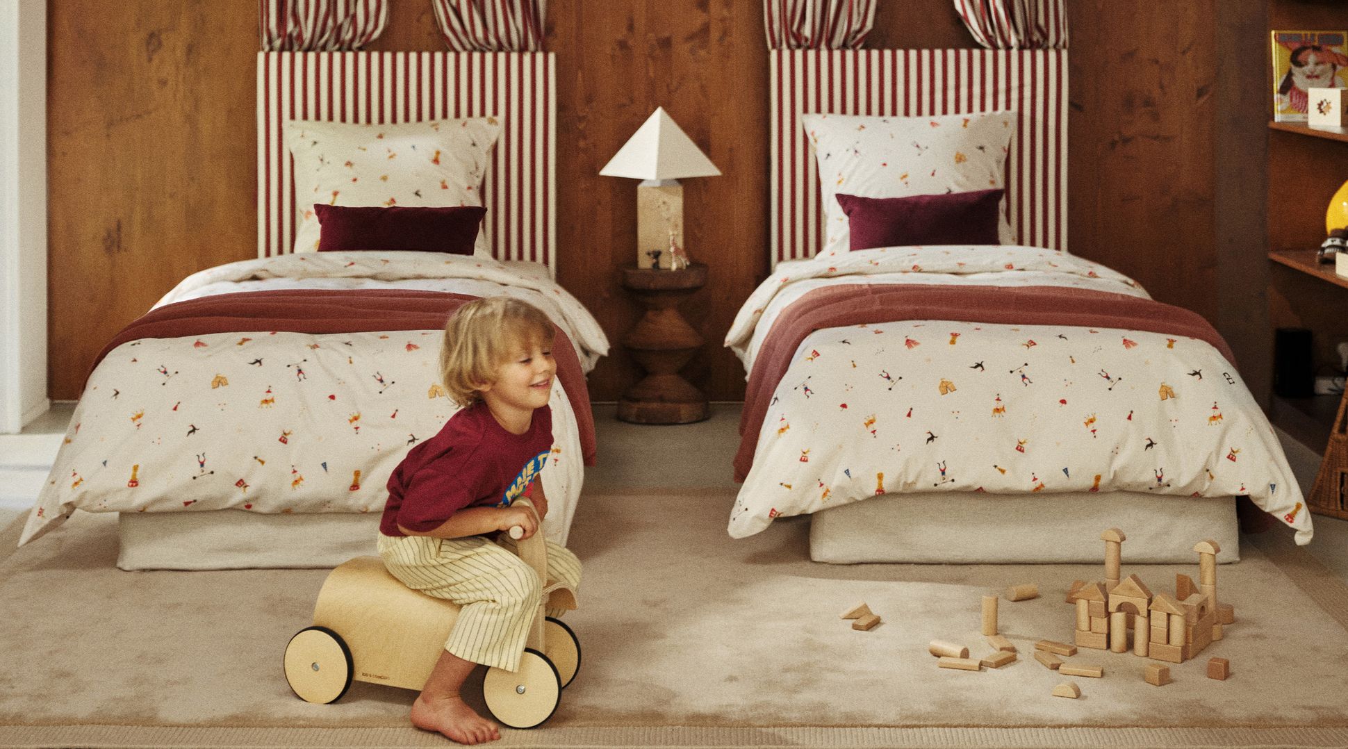 Un enfant joue avec un véhicule-jouet en bois dans une chambre chaleureuse dotée de deux lits, de têtes de lit à rayures, d'une literie rouge et blanche et de jouets en bois éparpillés sur le sol.