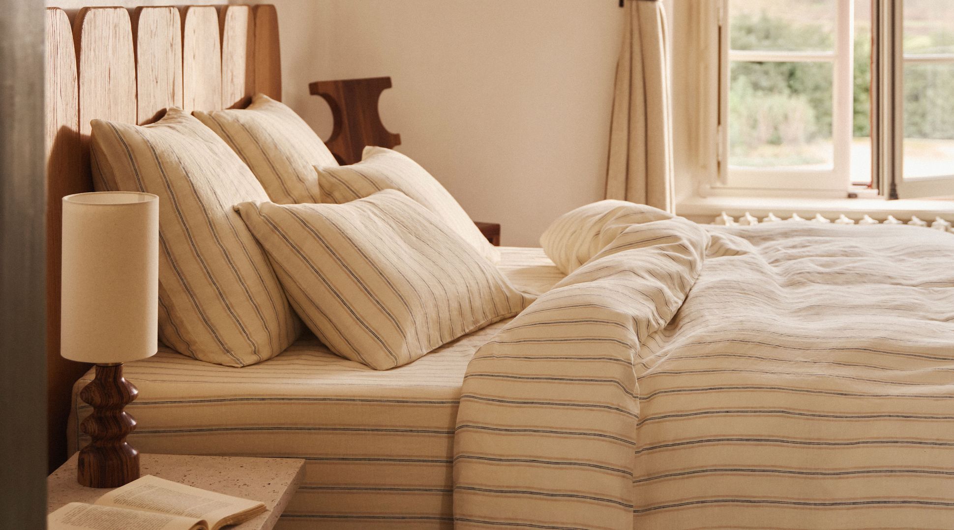 Une chambre douillette avec une tête de lit en bois, une parure de lit rayée, une lampe de chevet et un livre ouvert, baignée par la douce lumière naturelle qui pénètre par la fenêtre.