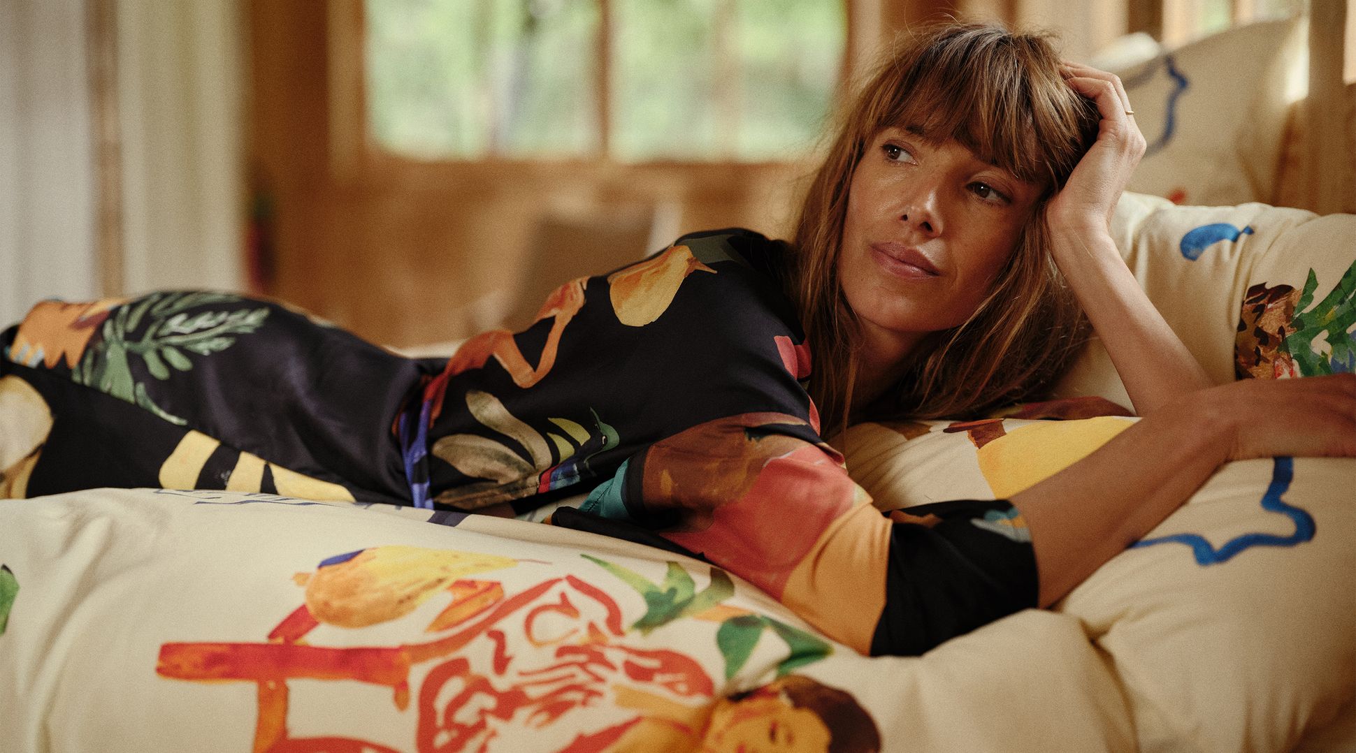 Une femme aux cheveux longs, vêtue d'une robe colorée, est allongée sur un coussin à motifs et regarde pensivement à travers une fenêtre floue en arrière-plan.