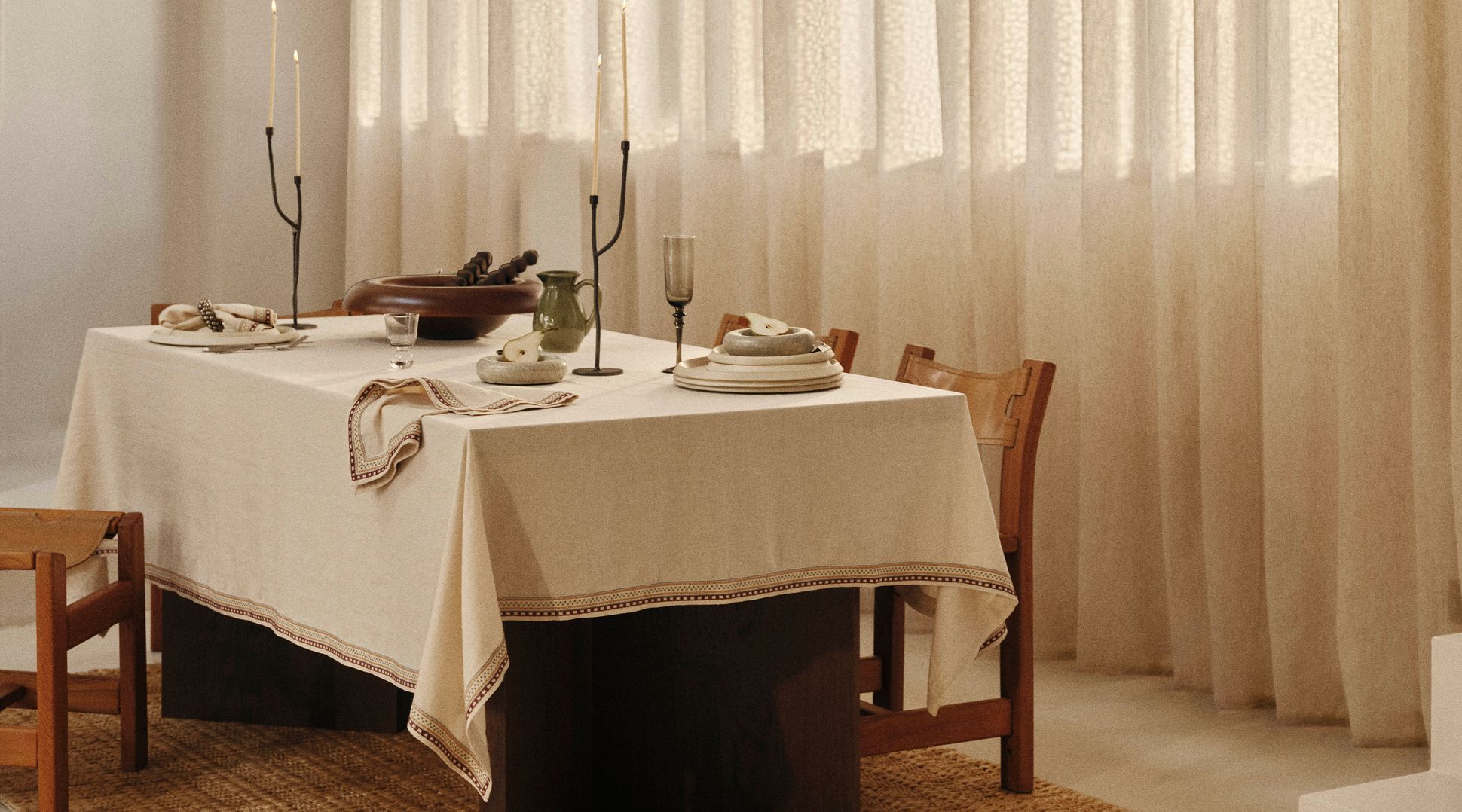 Une salle à manger minimaliste avec une table dressée pour deux, agrémentée de grandes bougies, d'une vaisselle sobre et de tons neutres, baignée d'une douce lumière naturelle.