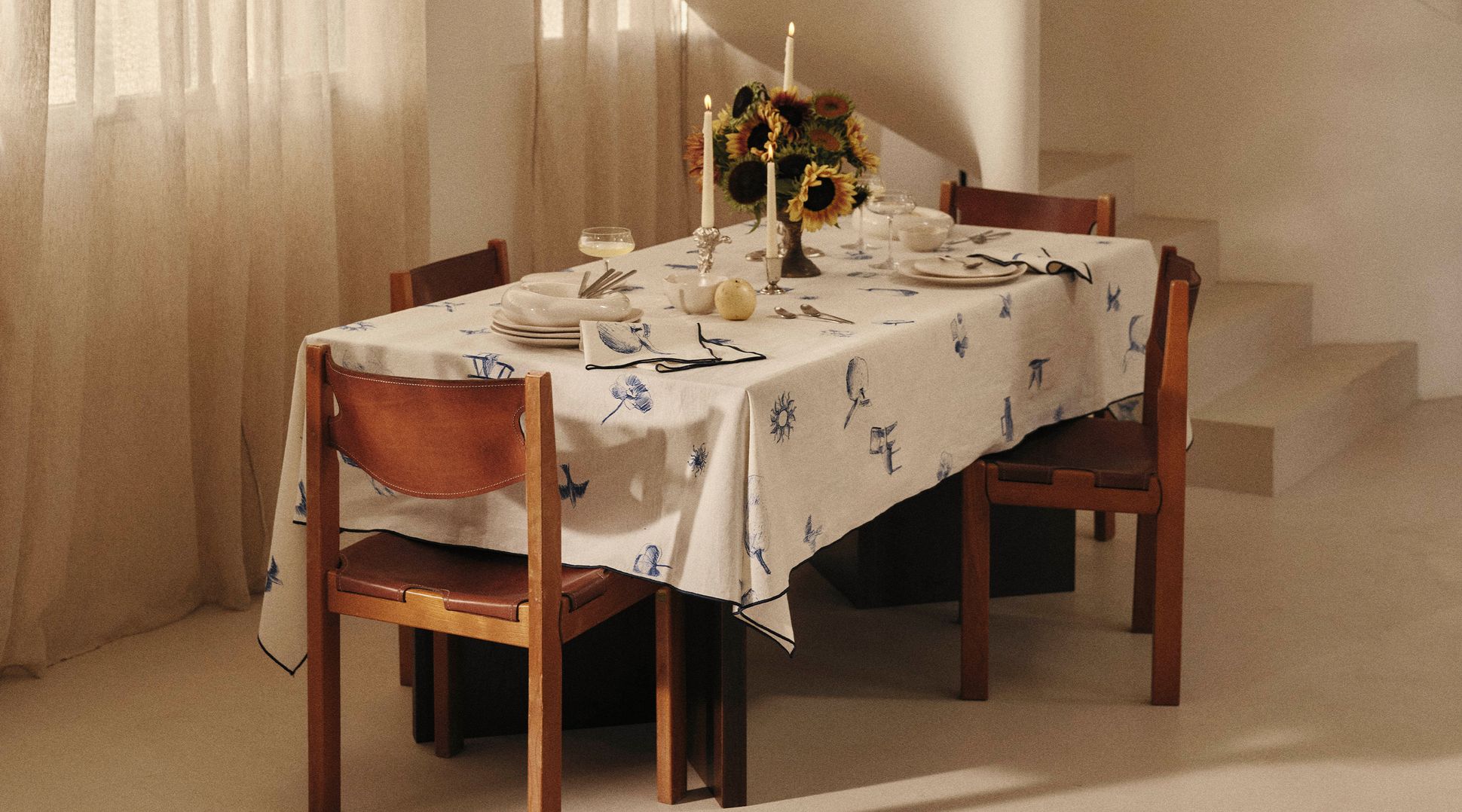 Une table à manger dressée pour quatre personnes, avec une nappe à motifs, des bougies et des tournesols, à côté d'un escalier en colimaçon et de rideaux légers et vaporeux.