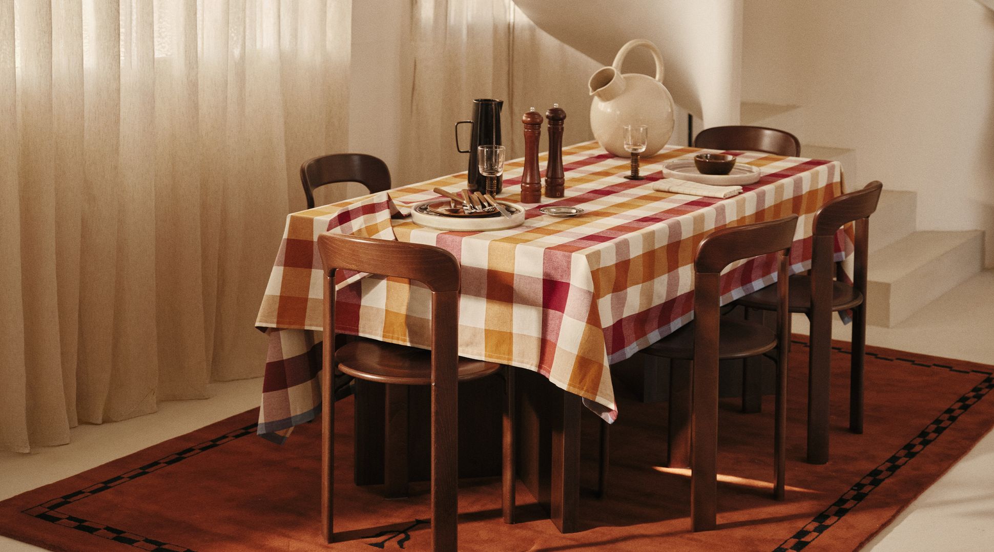 Salle à manger avec une table recouverte d'une nappe à carreaux, dressée avec une cruche, des salières et poivrières, et entourée de chaises en bois.