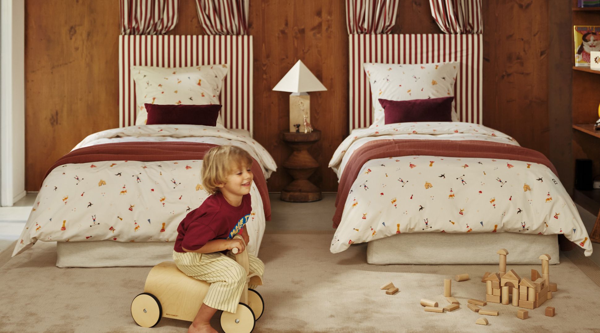 Un enfant joue avec un véhicule-jouet en bois dans une chambre chaleureuse dotée de deux lits, de têtes de lit à rayures, d'une literie rouge et blanche et de jouets en bois éparpillés sur le sol.