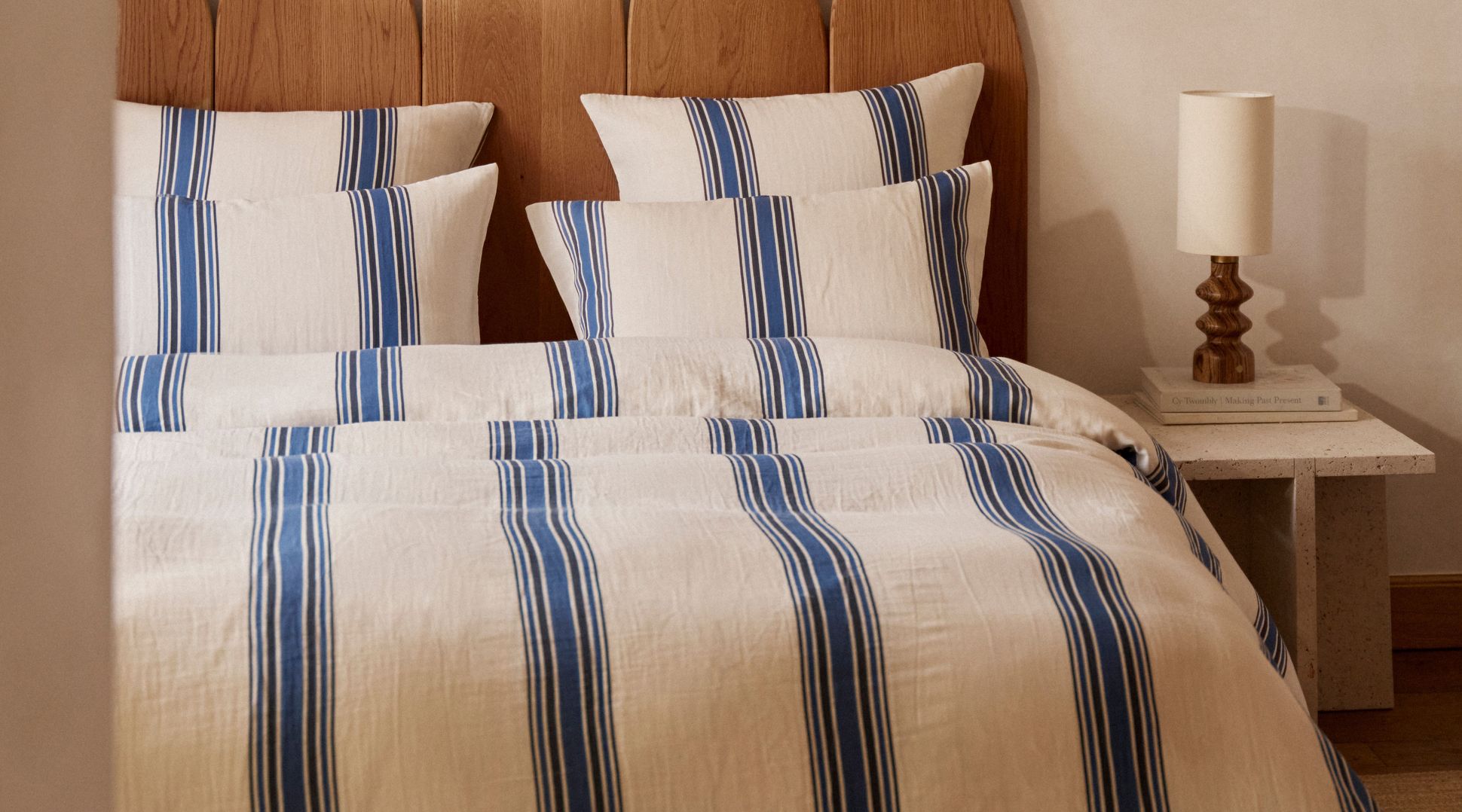 Un lit soigneusement fait, orné d'oreillers et d'une couette à rayures crème et bleues, avec une tête de lit en bois et une lampe posée sur une table de chevet.
