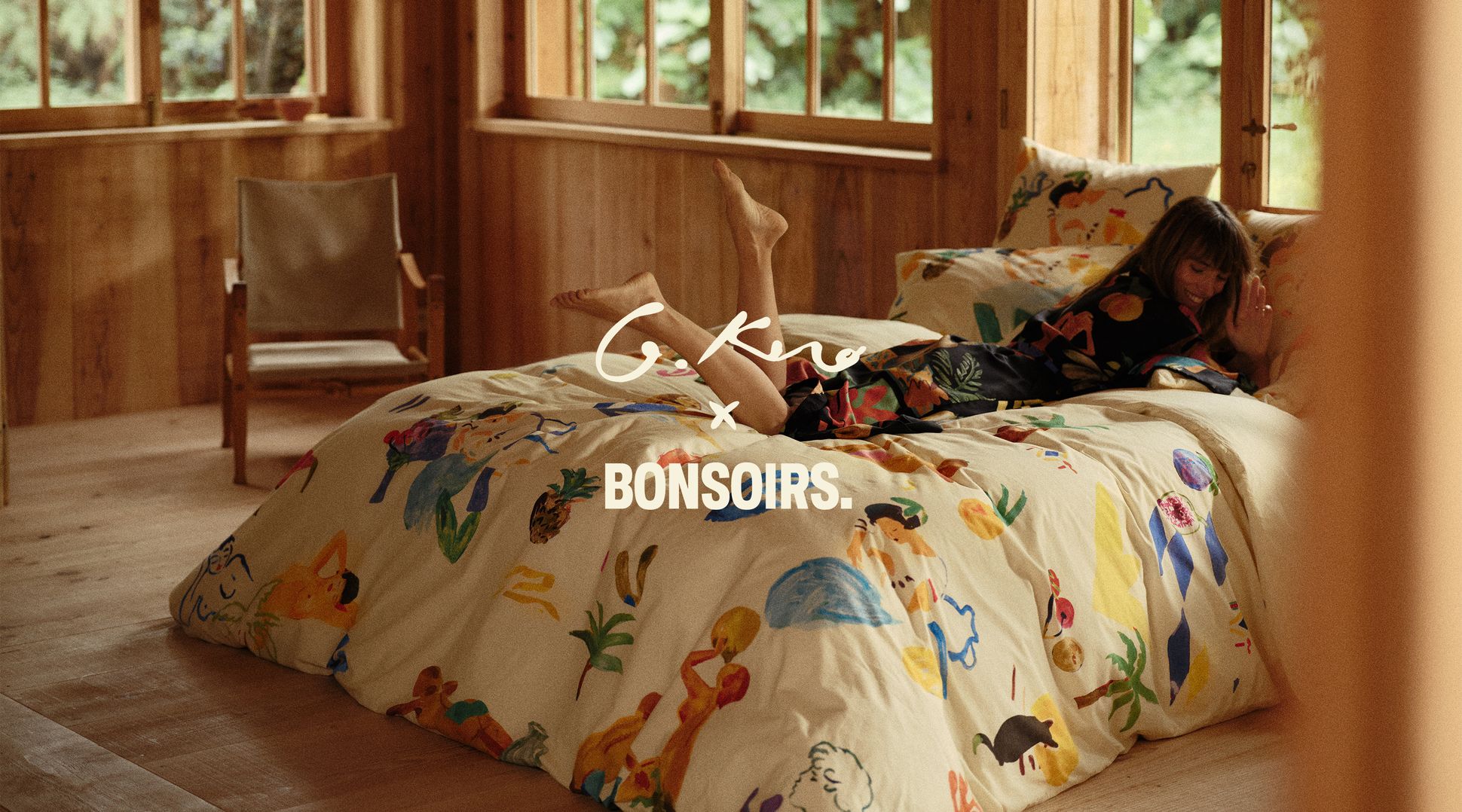 Une femme allongée sur un lit douillet aux draps colorés, dans une chambre aux murs en bois, avec de grandes fenêtres donnant sur la verdure ; on peut lire « G. Kero x Bonsoirs ».