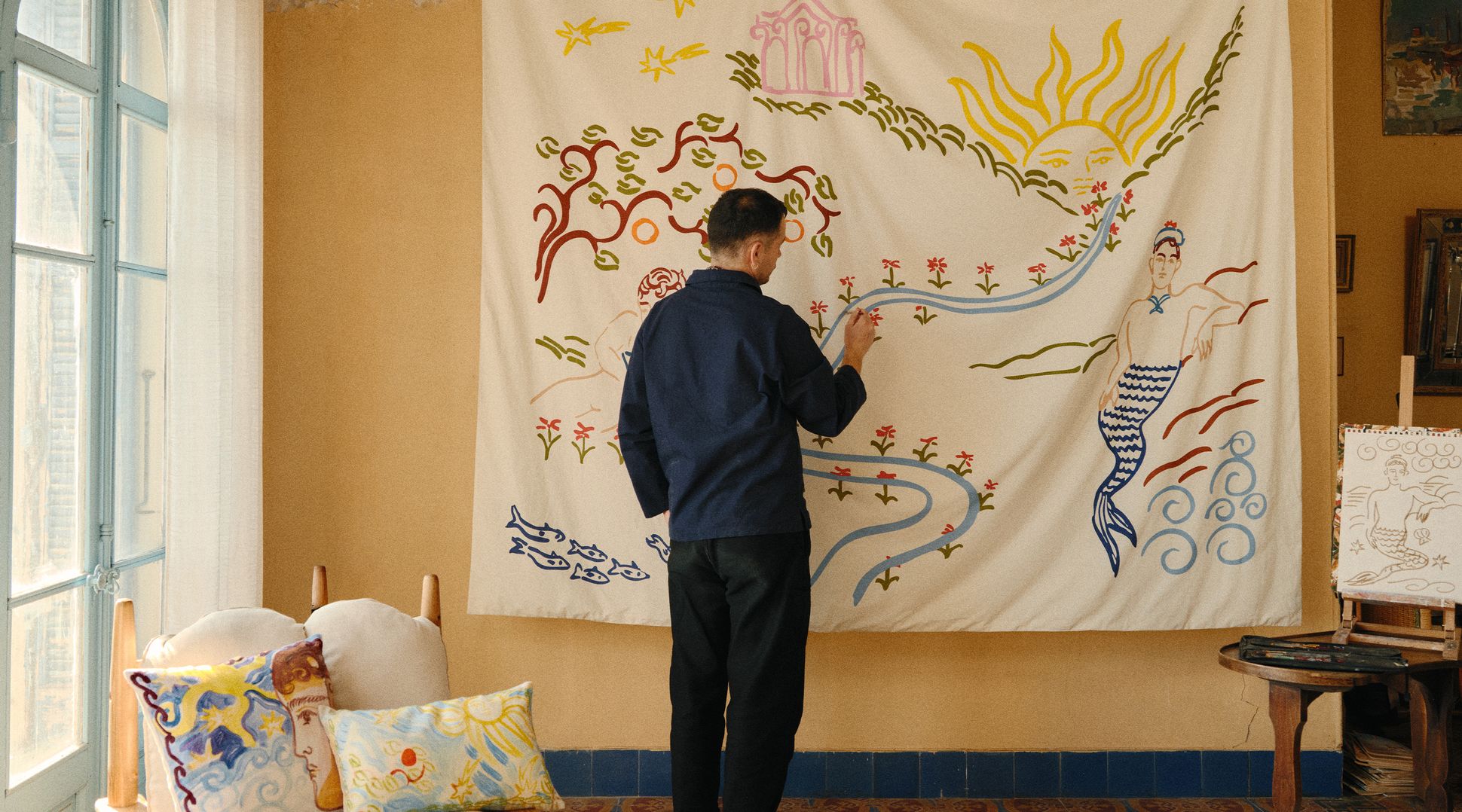 Une personne vêtue d'une chemise bleue peint sur une grande toile aux motifs fantaisistes, dans une pièce au sol à motifs et à la décoration vintage.