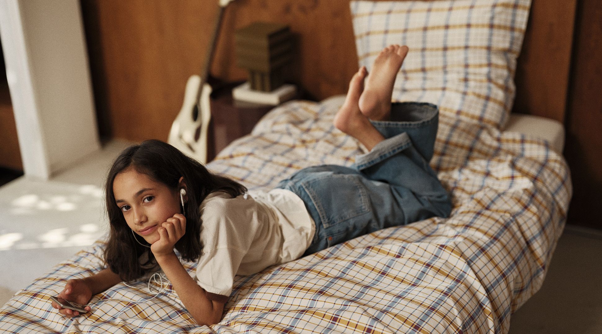 Une jeune fille allongée sur un lit, écoutant de la musique avec des écouteurs, un téléphone à la main. Le lit est recouvert d'une couverture à carreaux, et une douce lumière naturelle baigne la pièce.