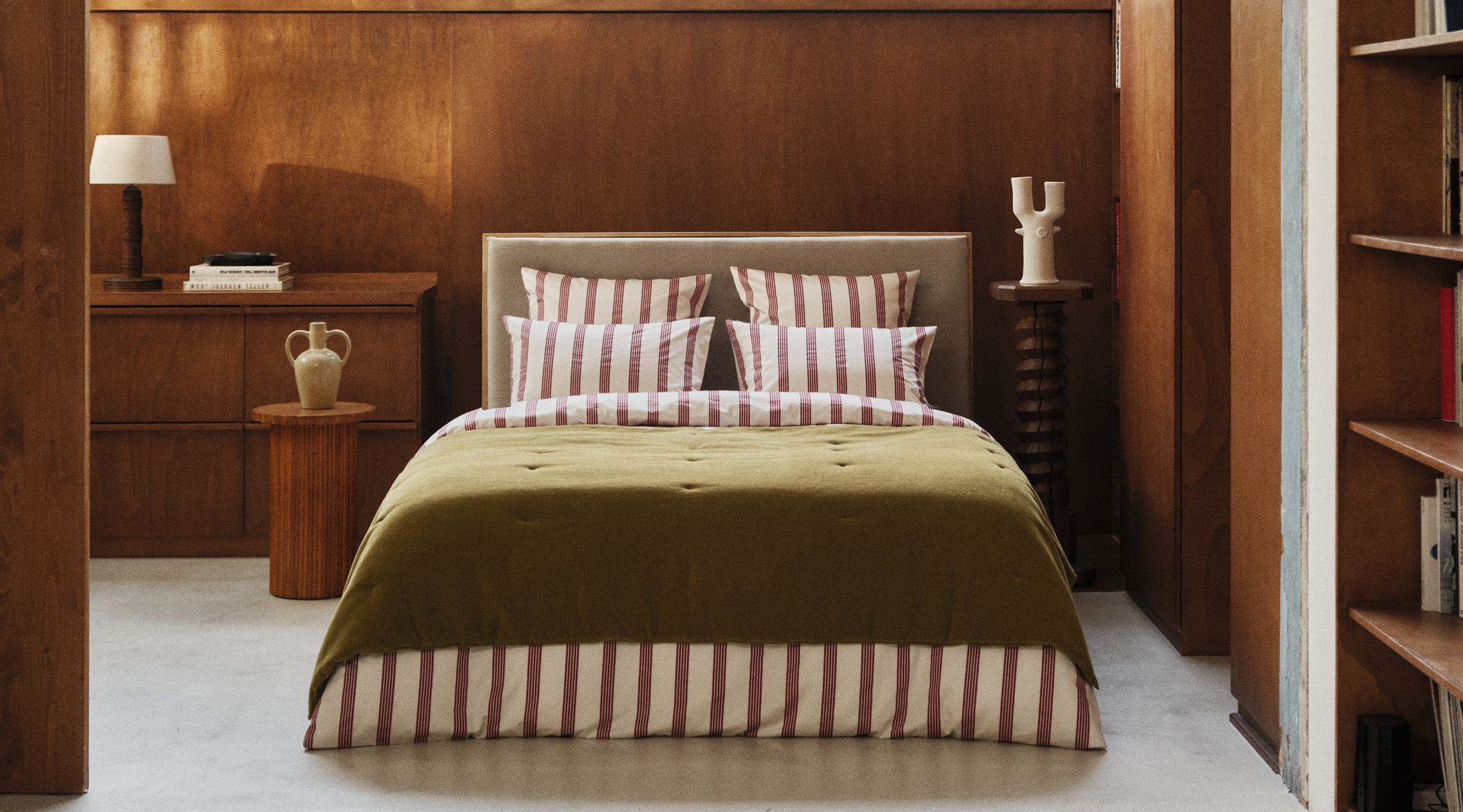Une chambre douillette avec un lit orné d'oreillers rayés et d'une couverture verte, des tables de chevet en bois, des vases, des livres et un éclairage tamisé, le tout devant un mur en bois.