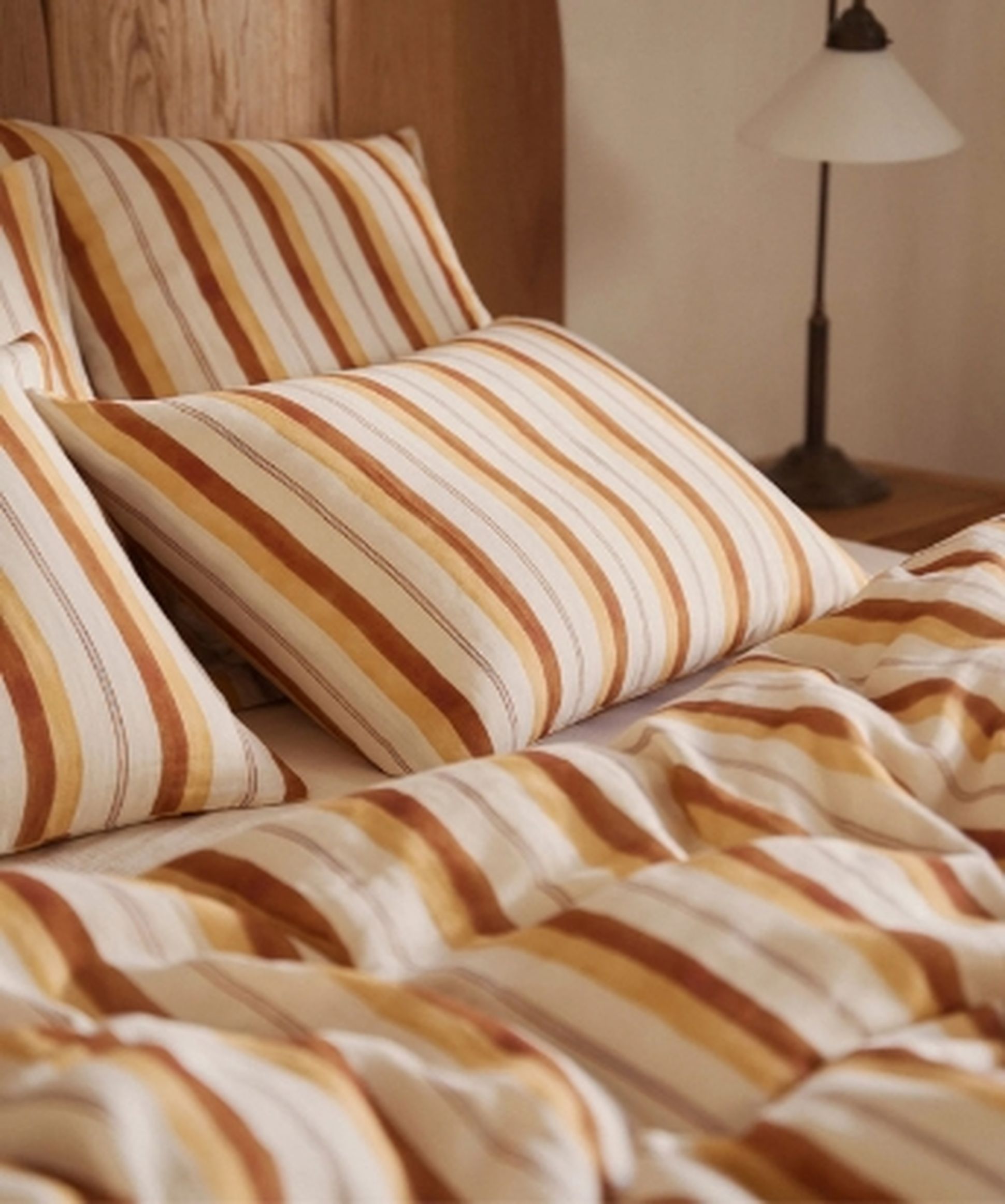 Un lit soigneusement fait, avec des oreillers et une couette à rayures alternées marron et blanches, à côté d'une tête de lit en bois et d'une lampe de chevet.