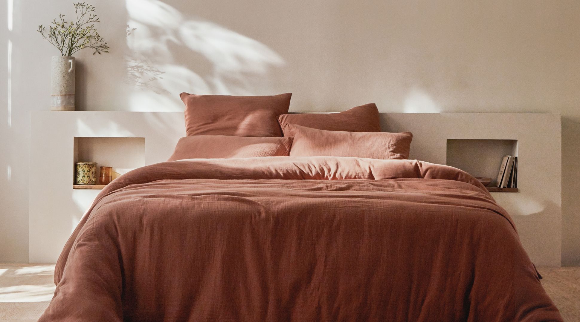 Un lit douillet avec une literie couleur rouille, adossé à un mur beige avec des étagères encastrées, et un vase avec des branches dessus.