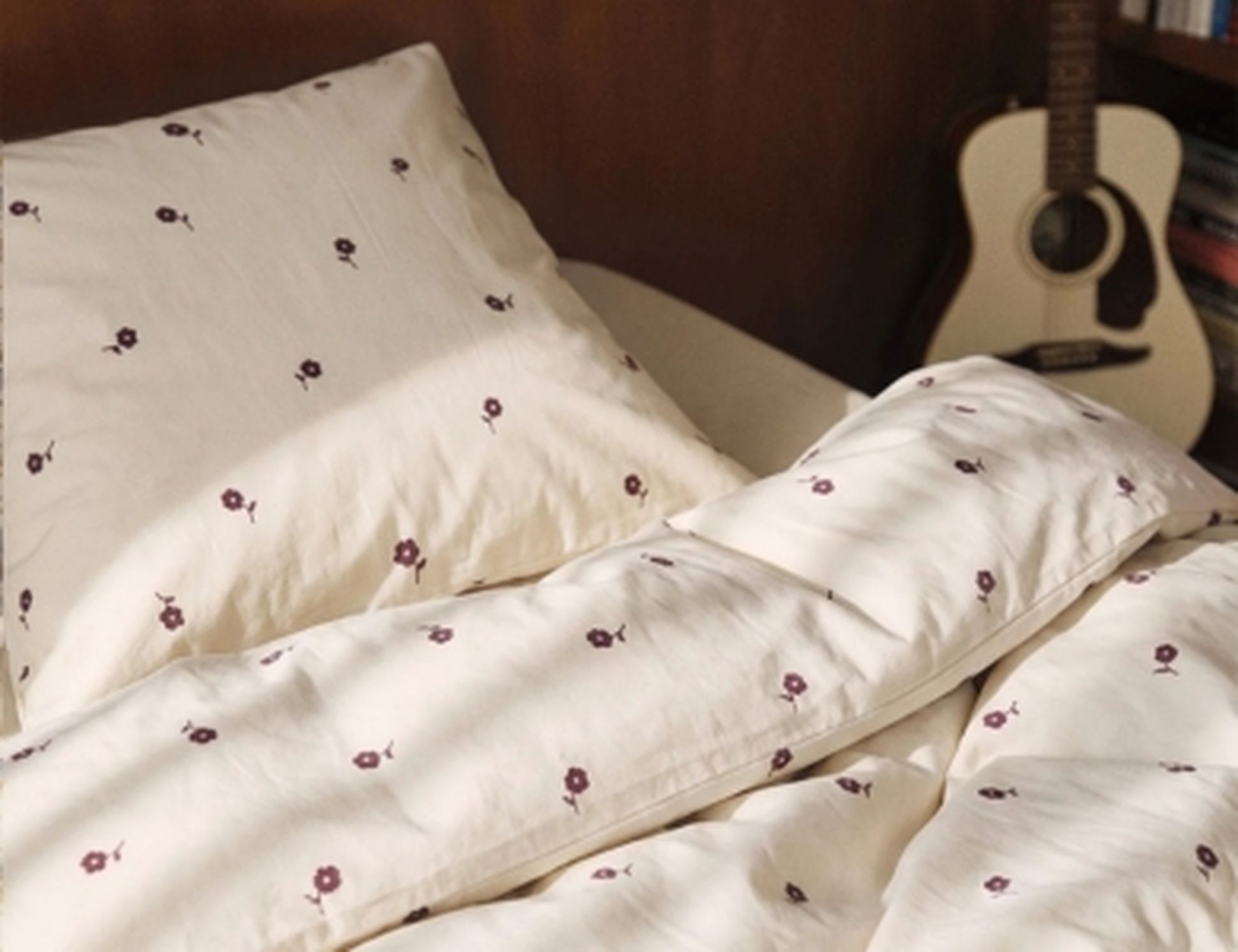 Un lit défait, avec des draps et des oreillers à motifs floraux, à côté d'une guitare acoustique, dans une chambre chaleureuse.