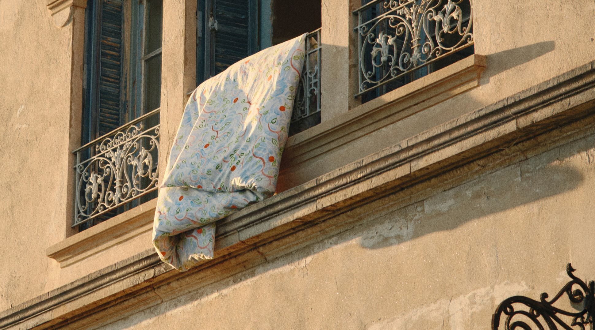 Une courtepointe colorée est suspendue à la balustrade en fer forgé d'un balcon, sur un immeuble aux fenêtres cintrées et aux volets bleus.