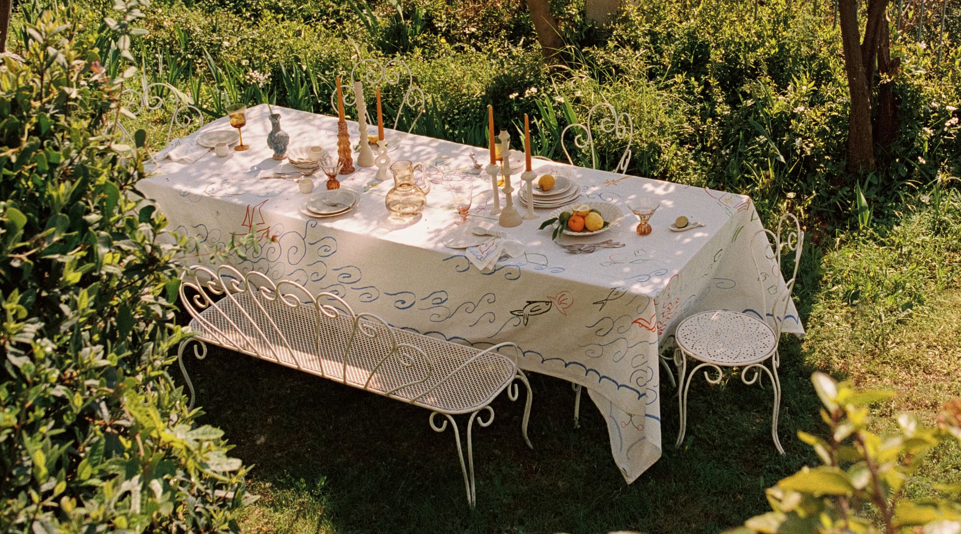 Une table d'extérieur dressée pour un repas, avec une nappe blanche, des bougies et des fruits, entourée d'une végétation luxuriante et à l'ombre d'un arbre.