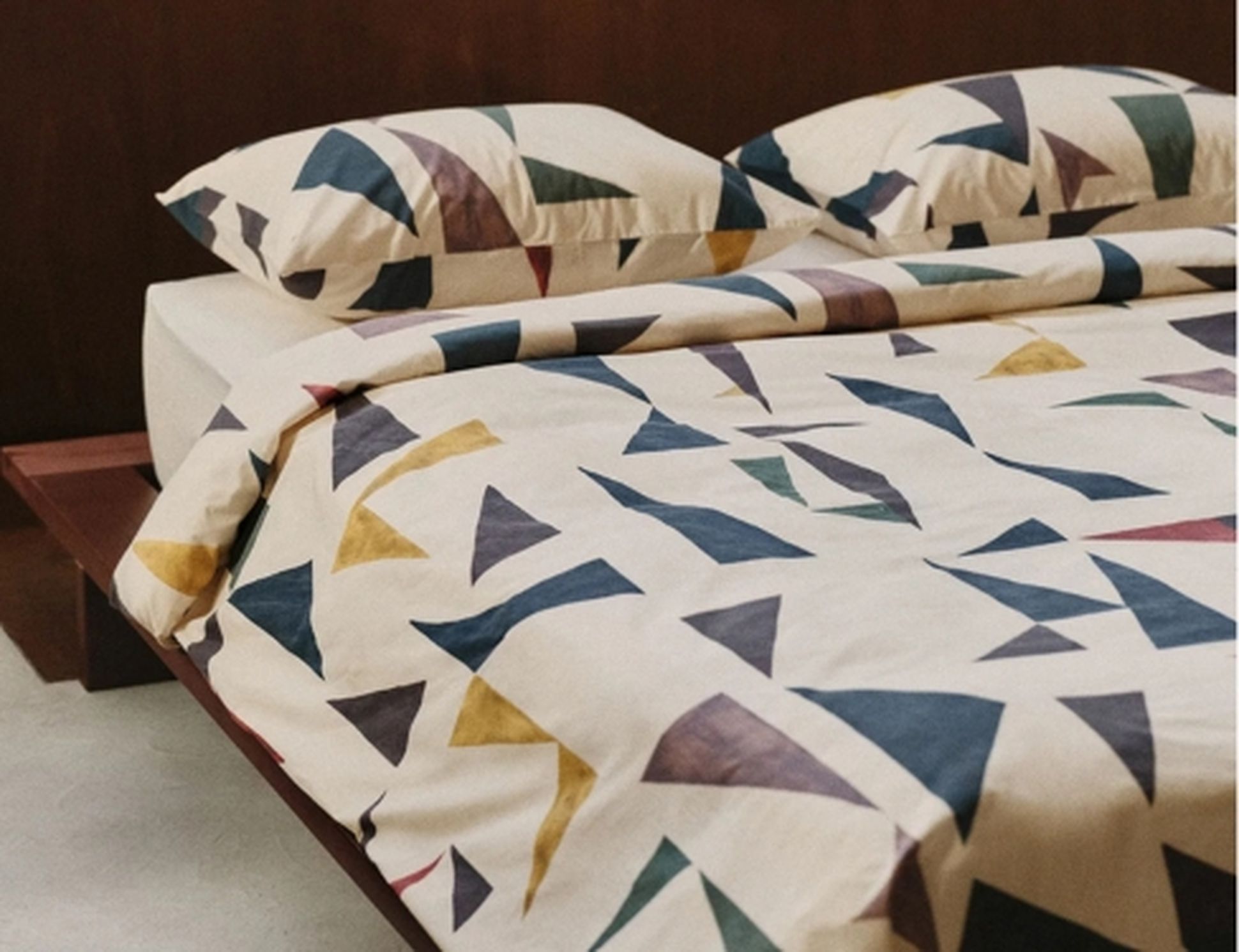 Lit avec draps et oreillers ornés d'un motif géométrique de triangles de différentes couleurs, dans une chambre minimaliste dotée d'une tête de lit en bois.