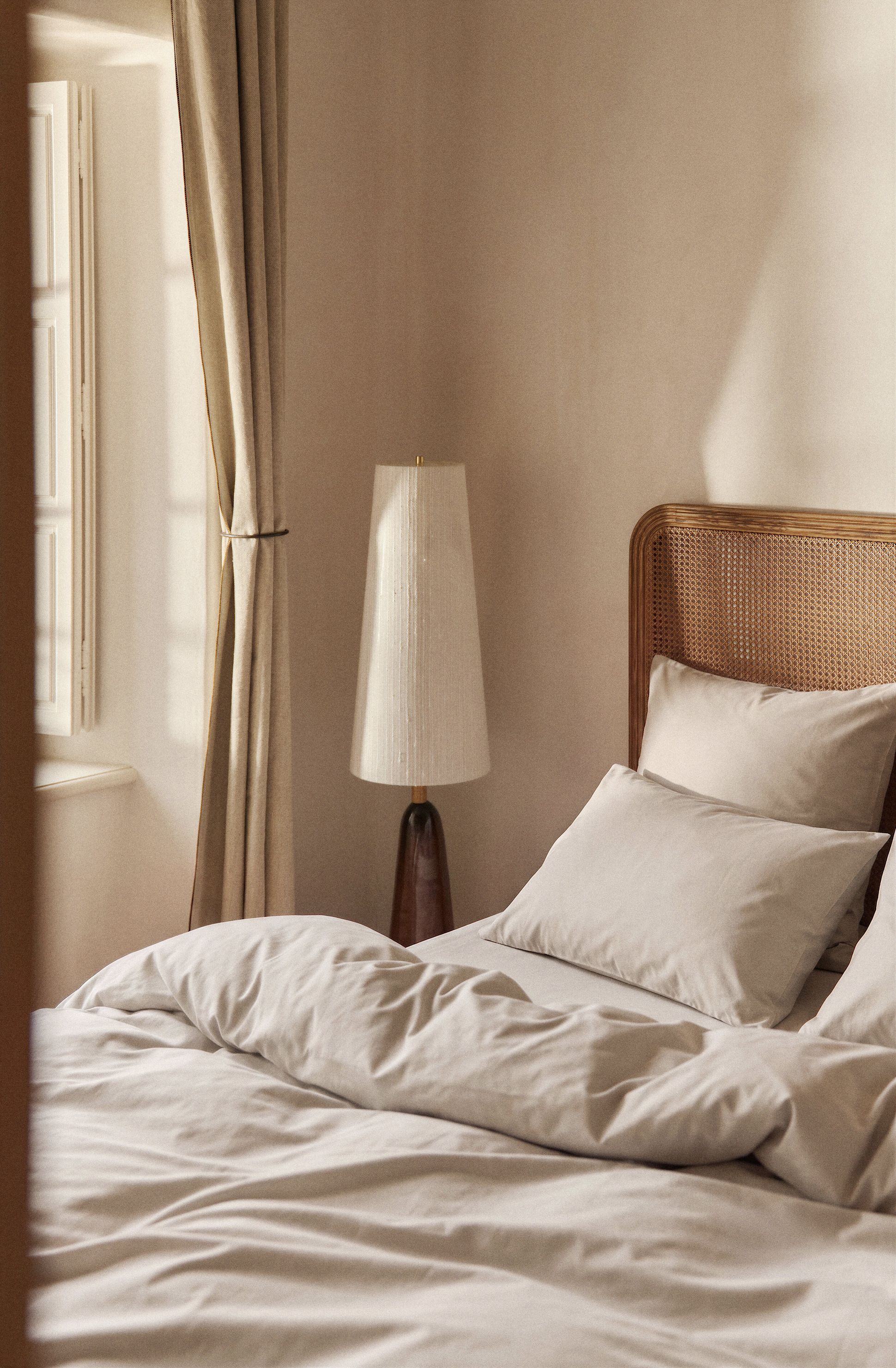 A cozy bedroom with a neatly made bed, beige bedding, a wooden headboard, a tall lamp, and sunlight filtering through curtains.