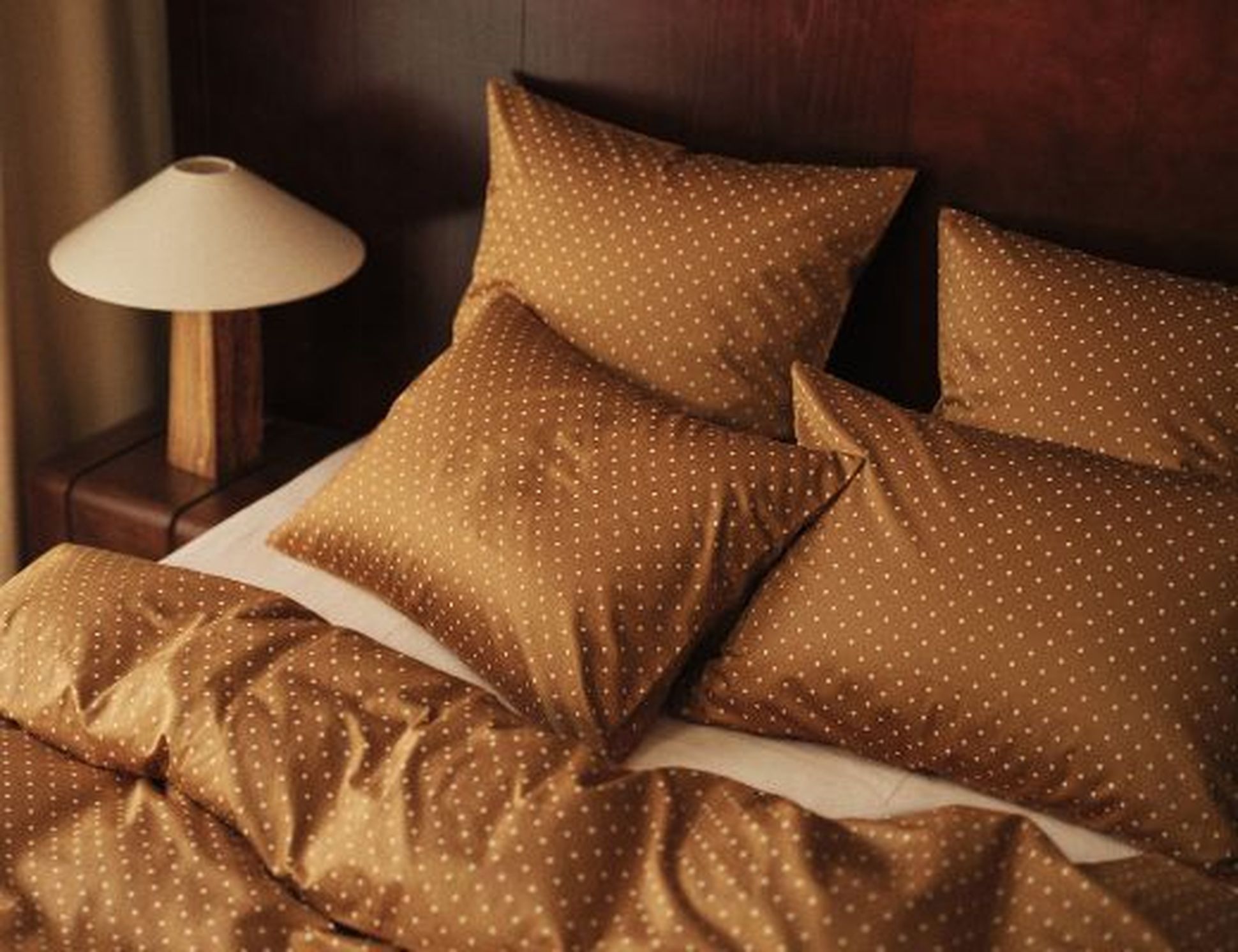 Lit doté de draps et d’oreillers à pois marron, placé à côté d’une table de chevet en bois surmontée d’une lampe, le tout adossé à une tête de lit en bois foncé.