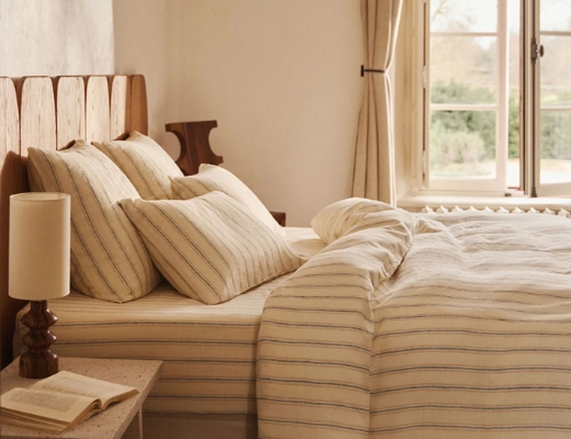 Une chambre douillette avec un lit bien fait, une parure de lit rayée et des oreillers moelleux près de la fenêtre. Sur la table de chevet se trouvent un livre et une lampe.