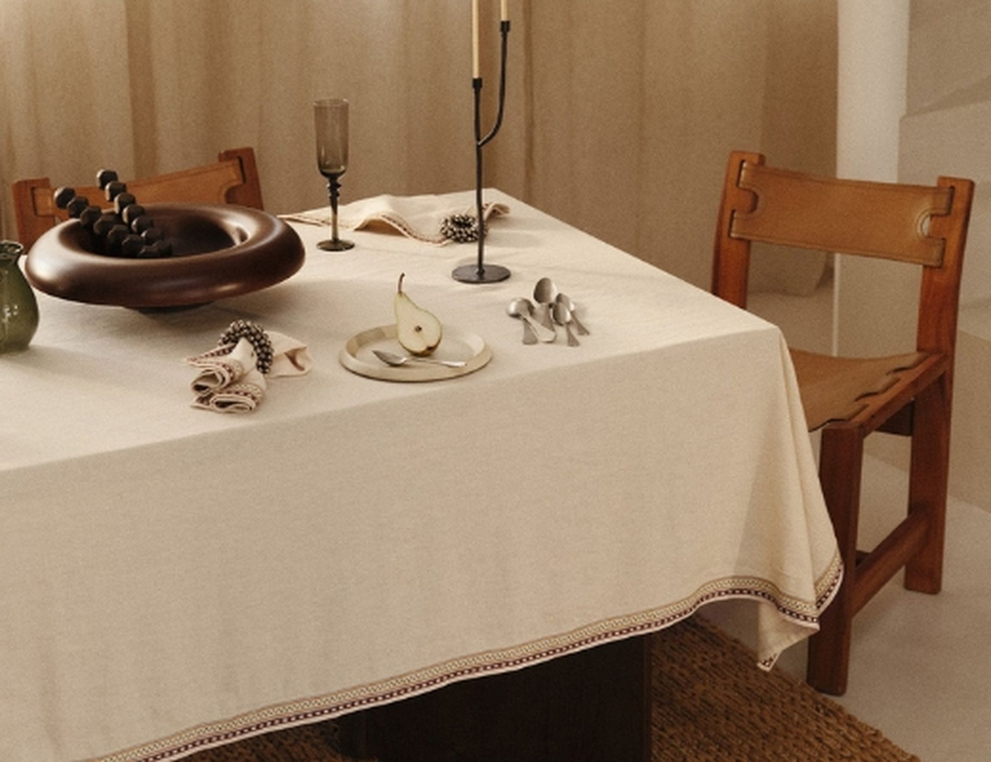 Une table à manger dressée avec une nappe blanche, une chaise marron, des assiettes ornées d'une tranche de poire, des couverts, un bol en bois, des bougeoirs et un verre à vin.