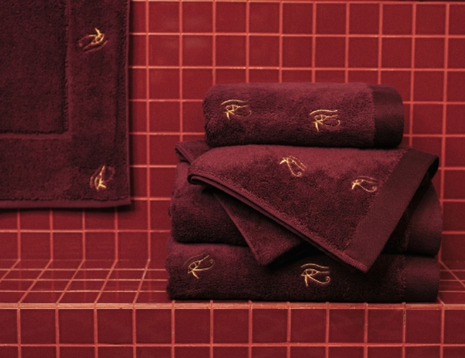 Une pile de serviettes bordeaux ornées de broderies dorées, posées sur un carrelage assorti.