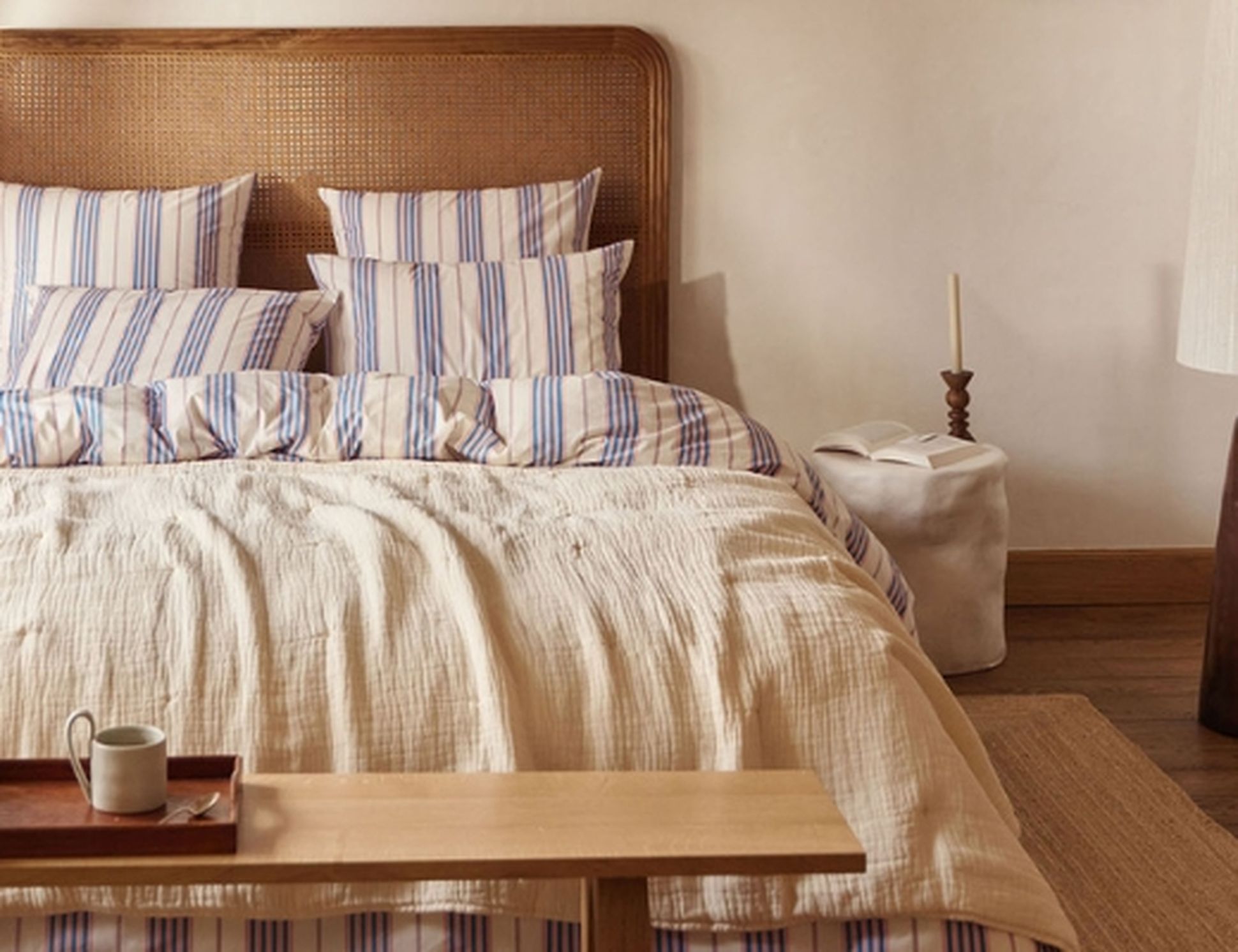 Chambre confortable avec une tête de lit en rotin, une literie rayée, un banc en bois et une décoration minimaliste comprenant une bougie et un livre ouvert.
